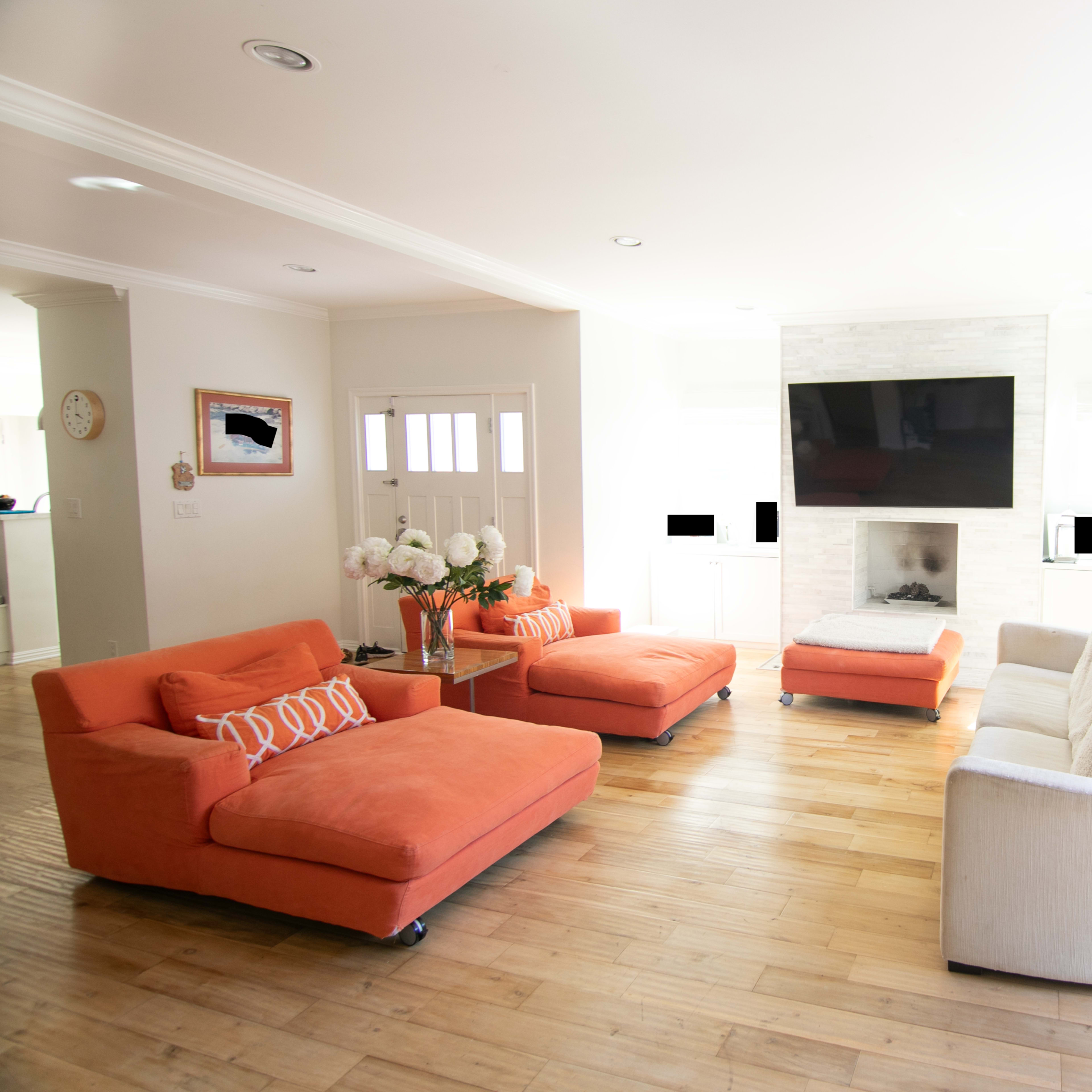 A bright living room featuring two orange lounge chairs, a light-colored sofa, a large flat-screen TV mounted above a fireplace, and a sliding door leading to an outdoor area.