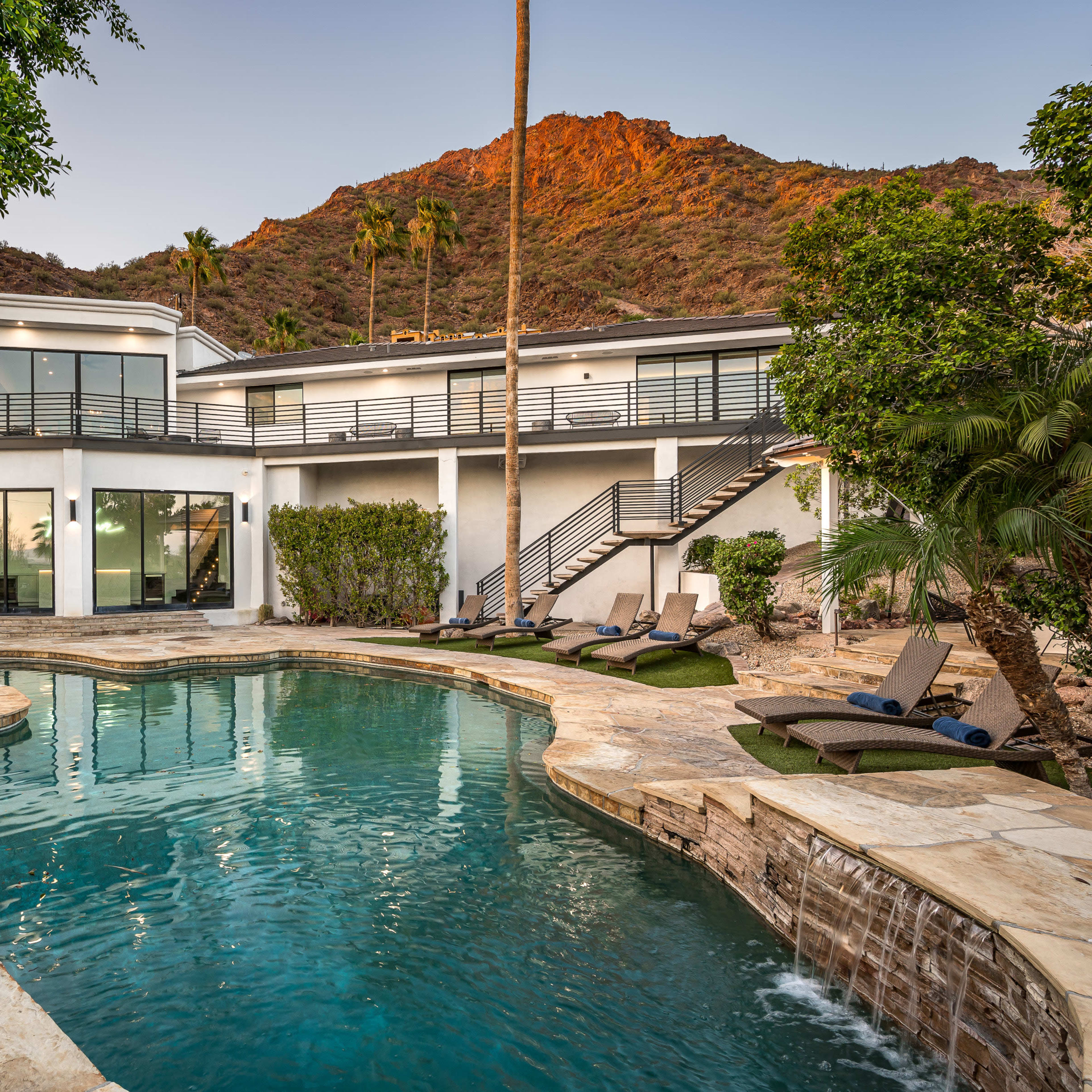 A modern home with a swimming pool surrounded by stone decking and palm trees, set against a mountain backdrop.