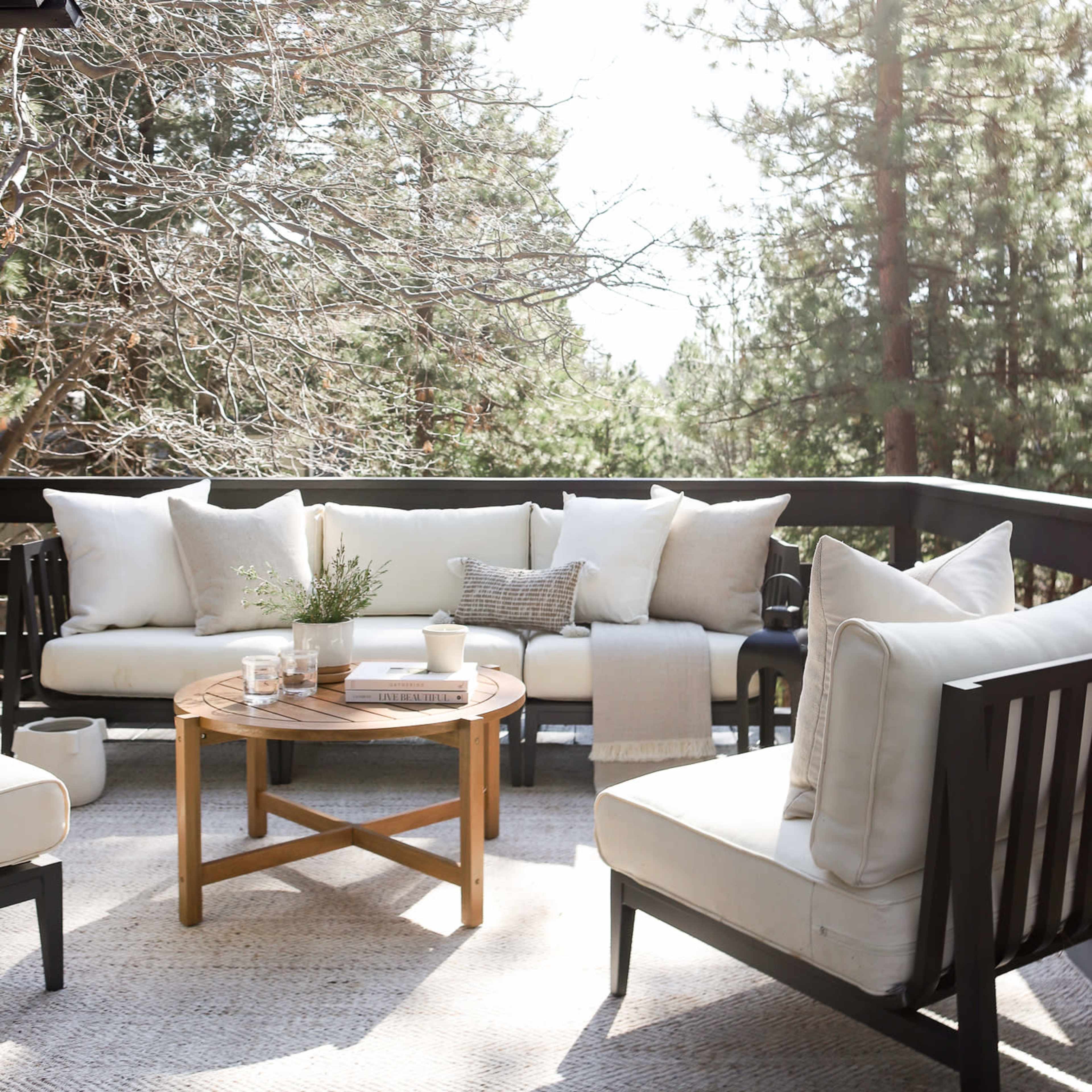 A light-filled outdoor patio features a cozy seating arrangement with neutral cushions, a round wooden coffee table, and a textured area rug surrounded by tall trees.