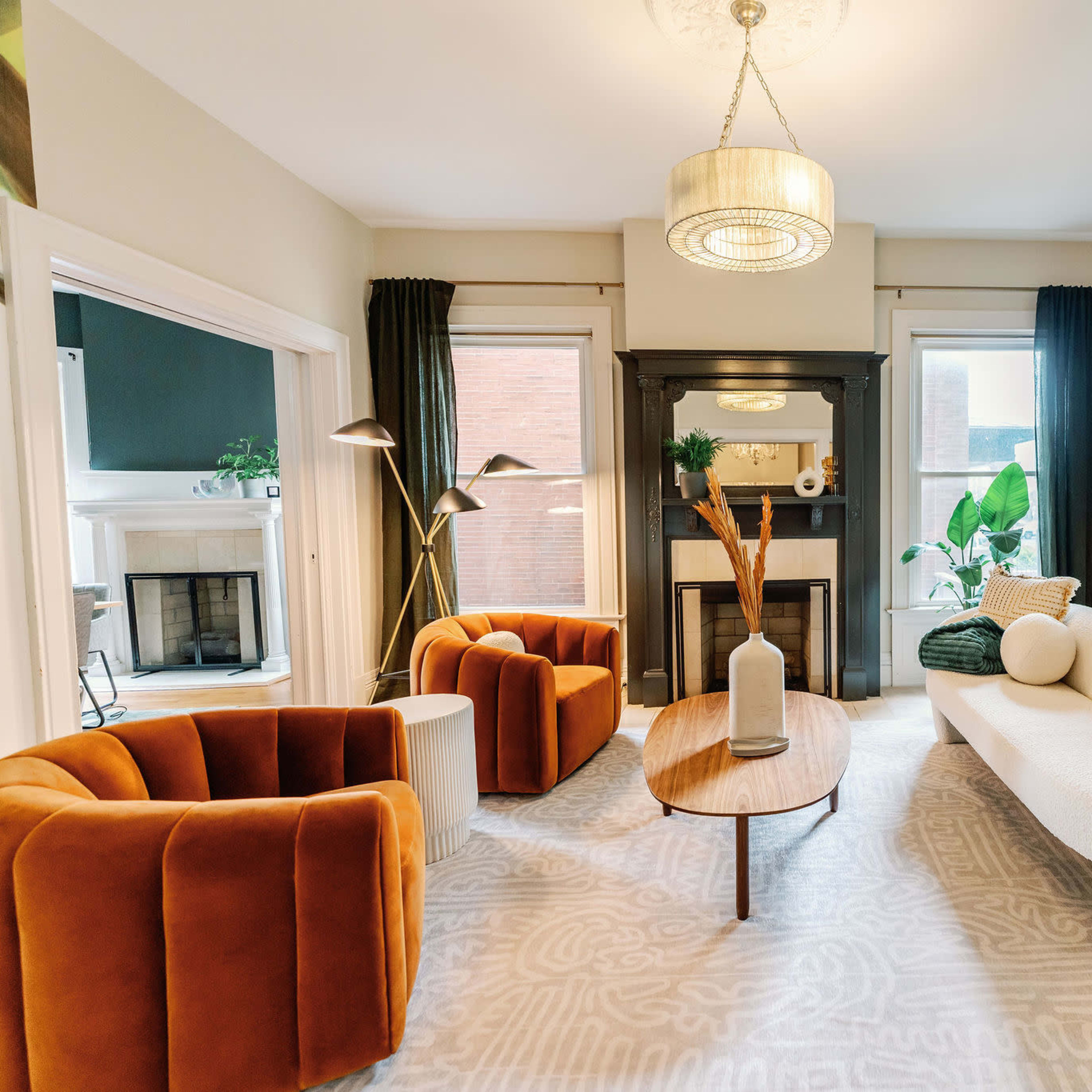 A modern living room features two orange armchairs, a white sofa with decorative pillows, a round wooden coffee table, and large windows with green drapes.