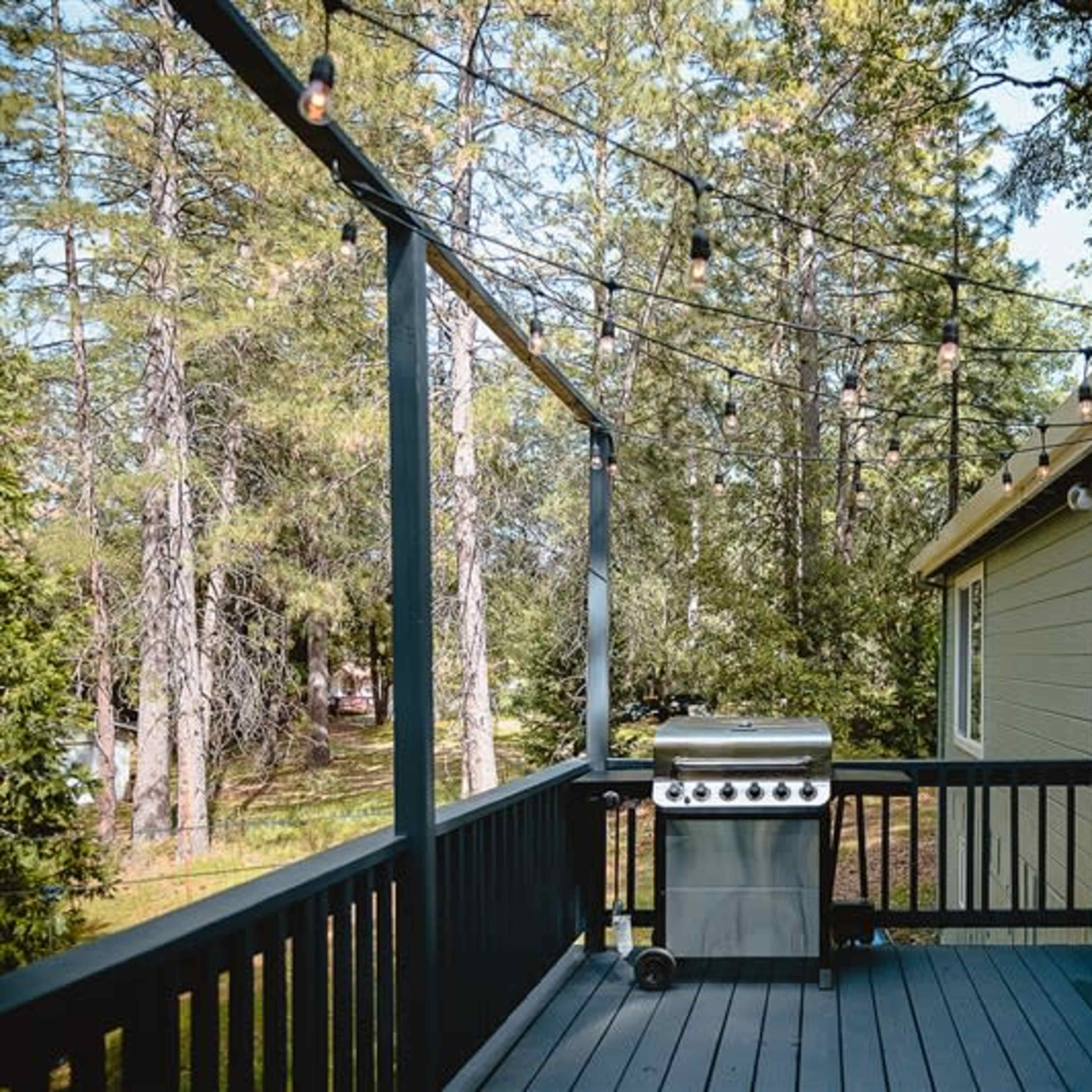 A wooden deck featuring a gas grill and string lights, surrounded by tall trees.
