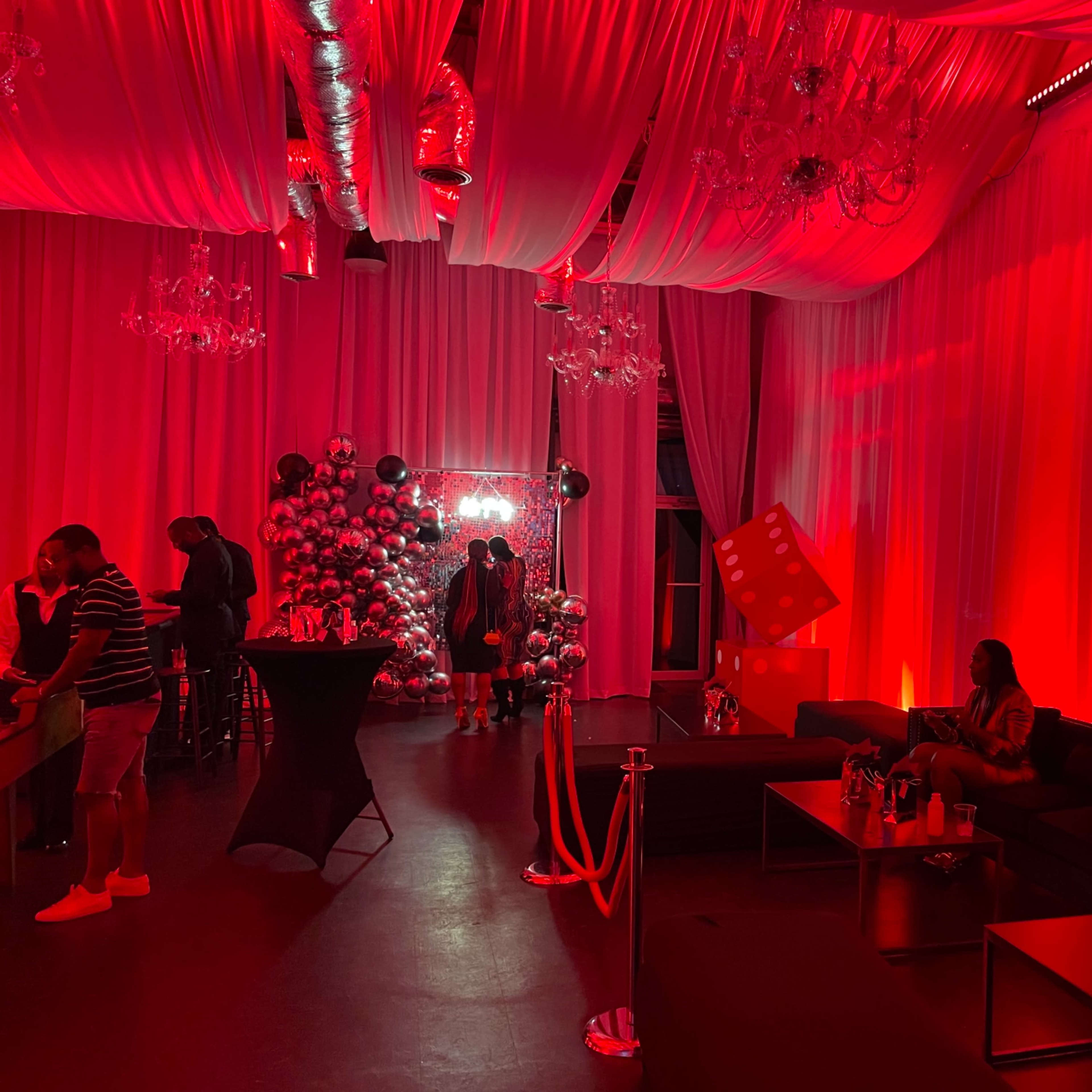The interior of a dimly lit event space is decorated with red lighting, featuring a Christmas tree and several seating areas.
