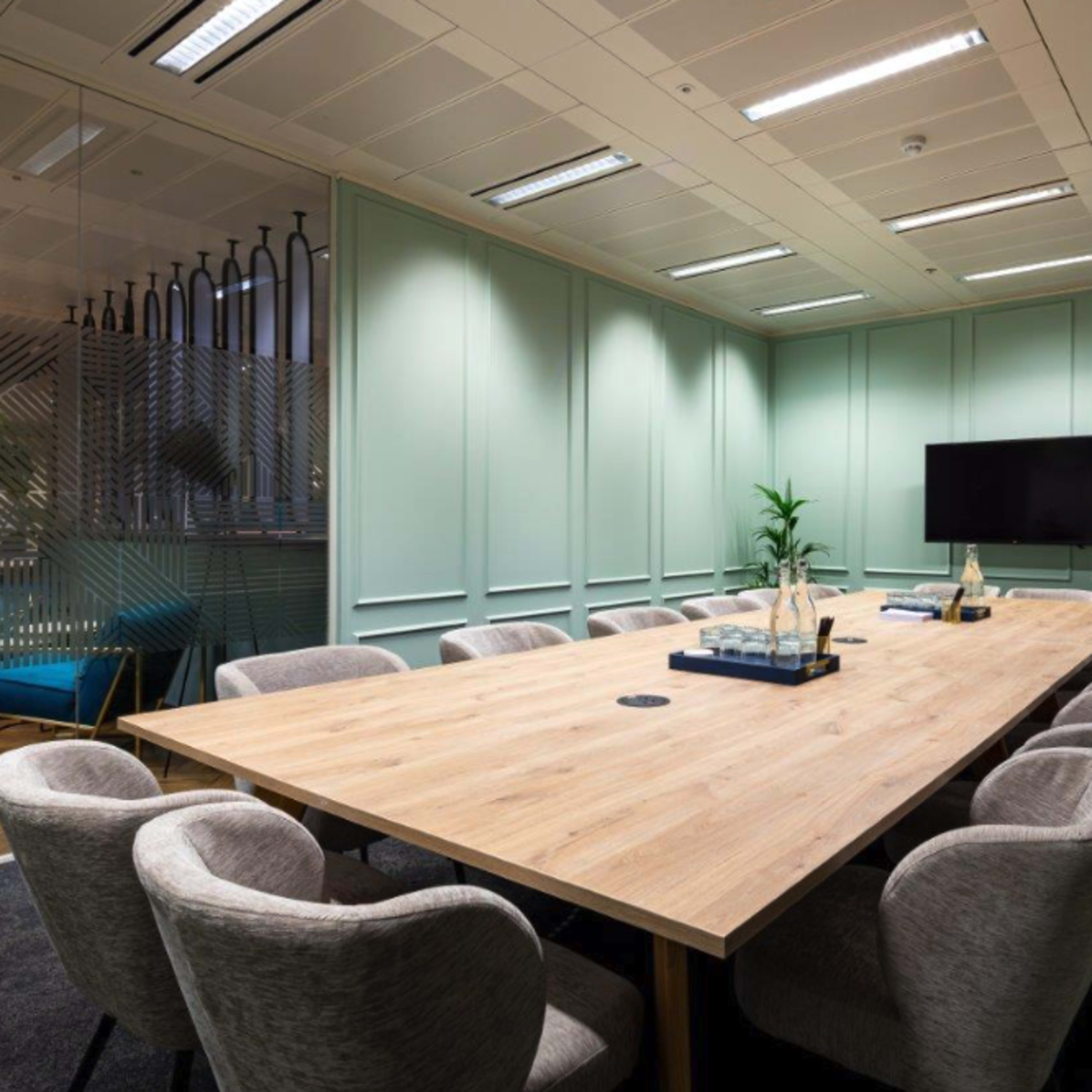 A modern conference room features a large wooden table surrounded by plush gray chairs, with green walls and a television on one side.