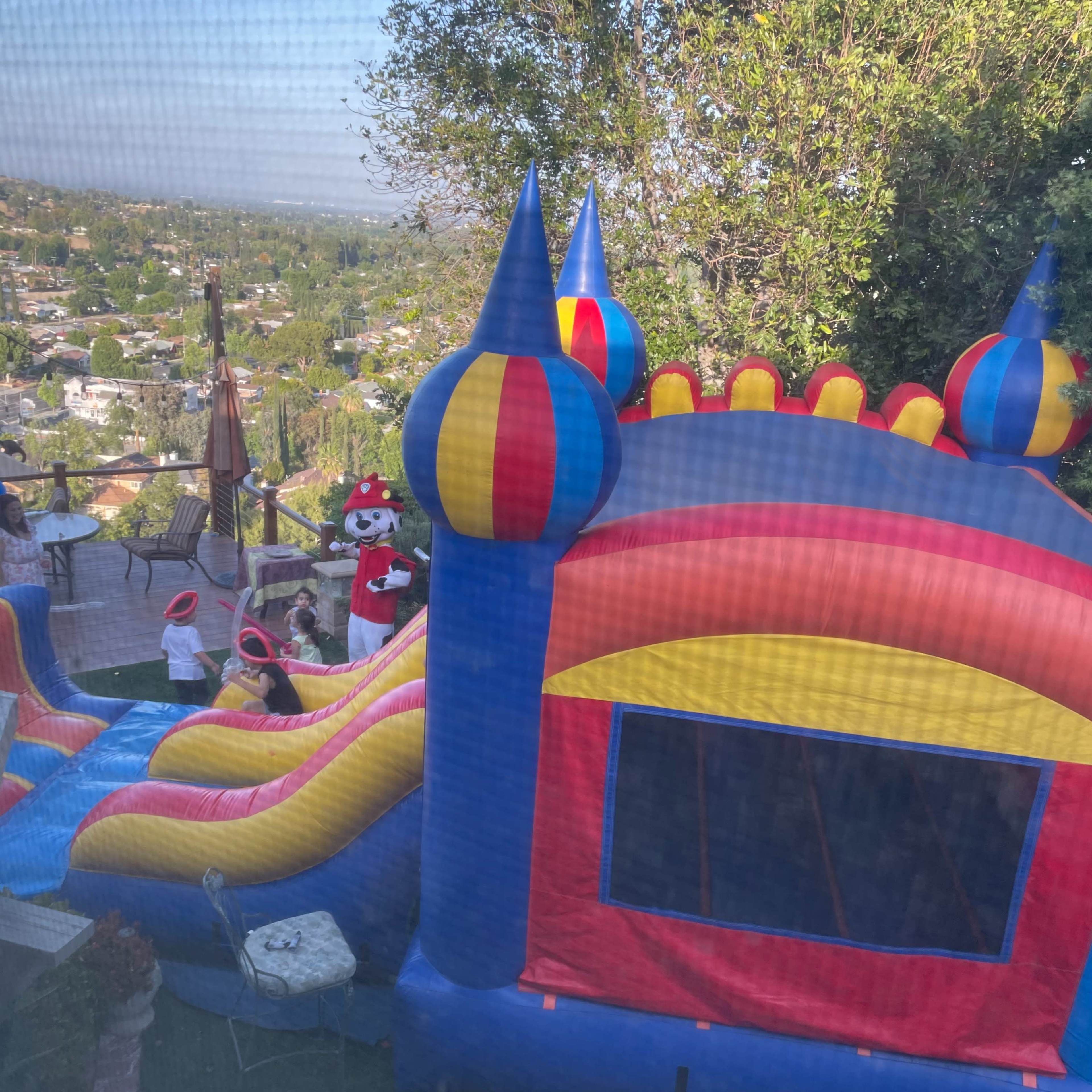 A colorful bounce house is set up in a backyard, with children playing nearby and a view of the neighborhood in the background.