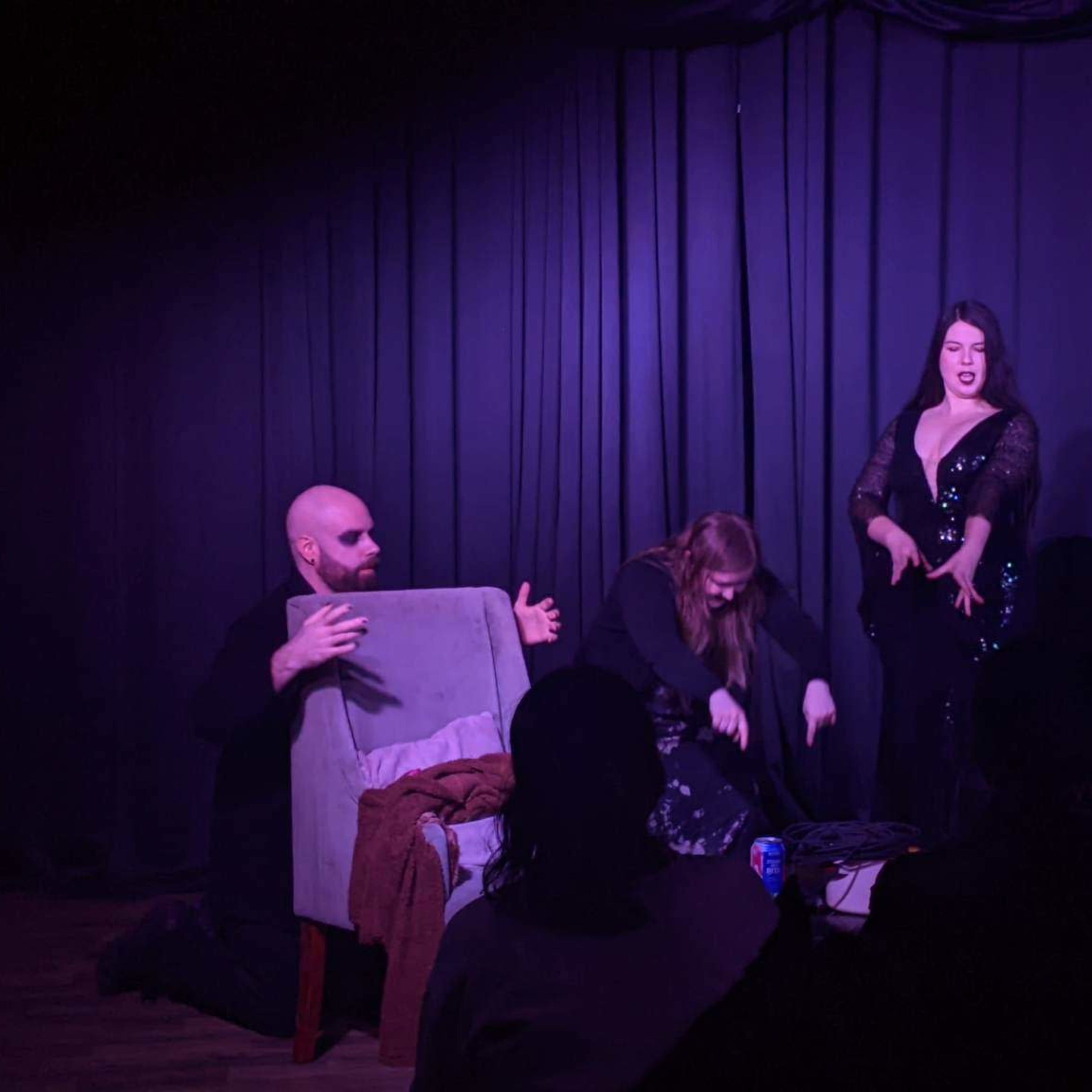 A performer in a black sequined outfit stands confidently on stage while two individuals interact with a chair in front of her.