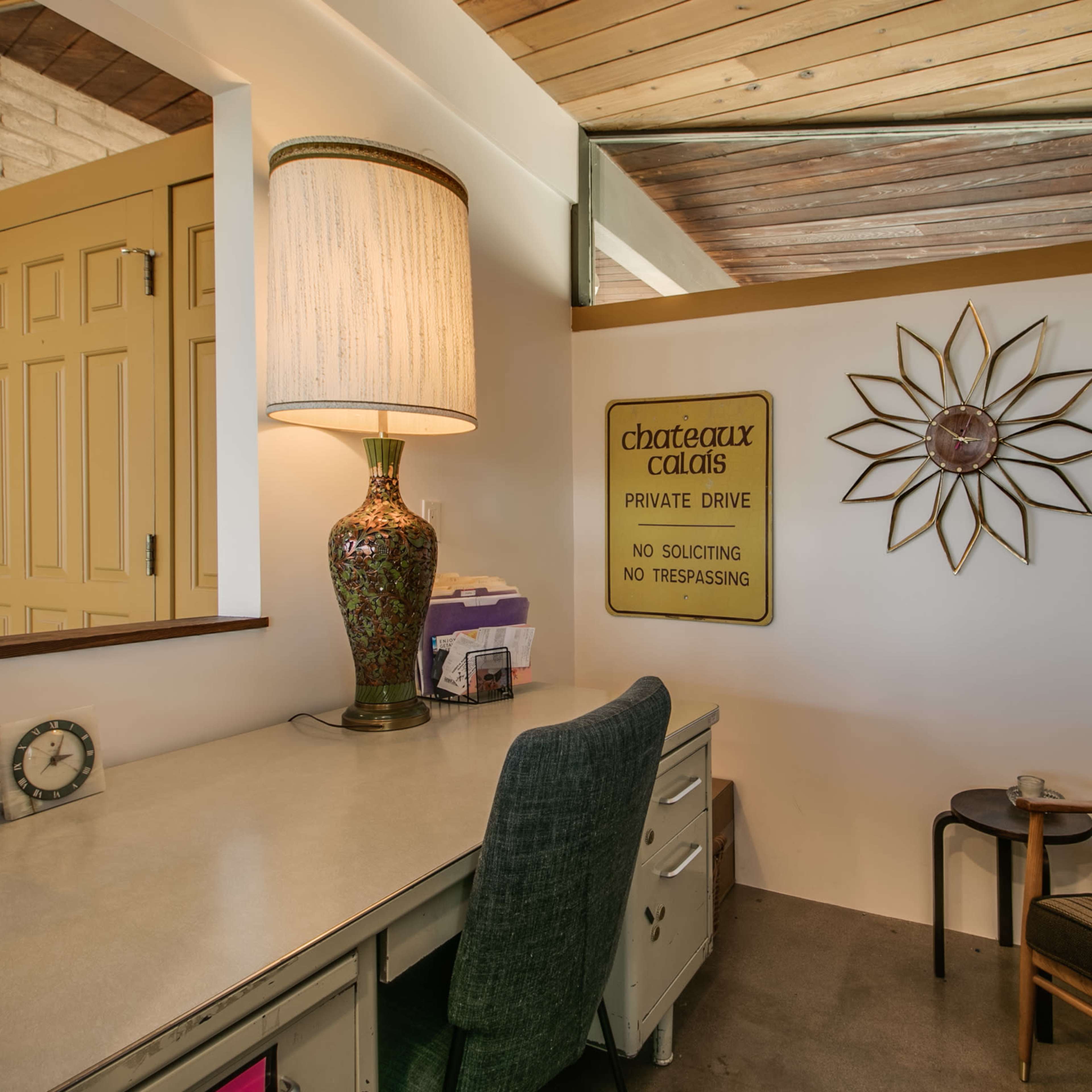 A cozy home office setup featuring a desk, a lamp, a decorative wall sign, and a chair against a backdrop of wooden beams.