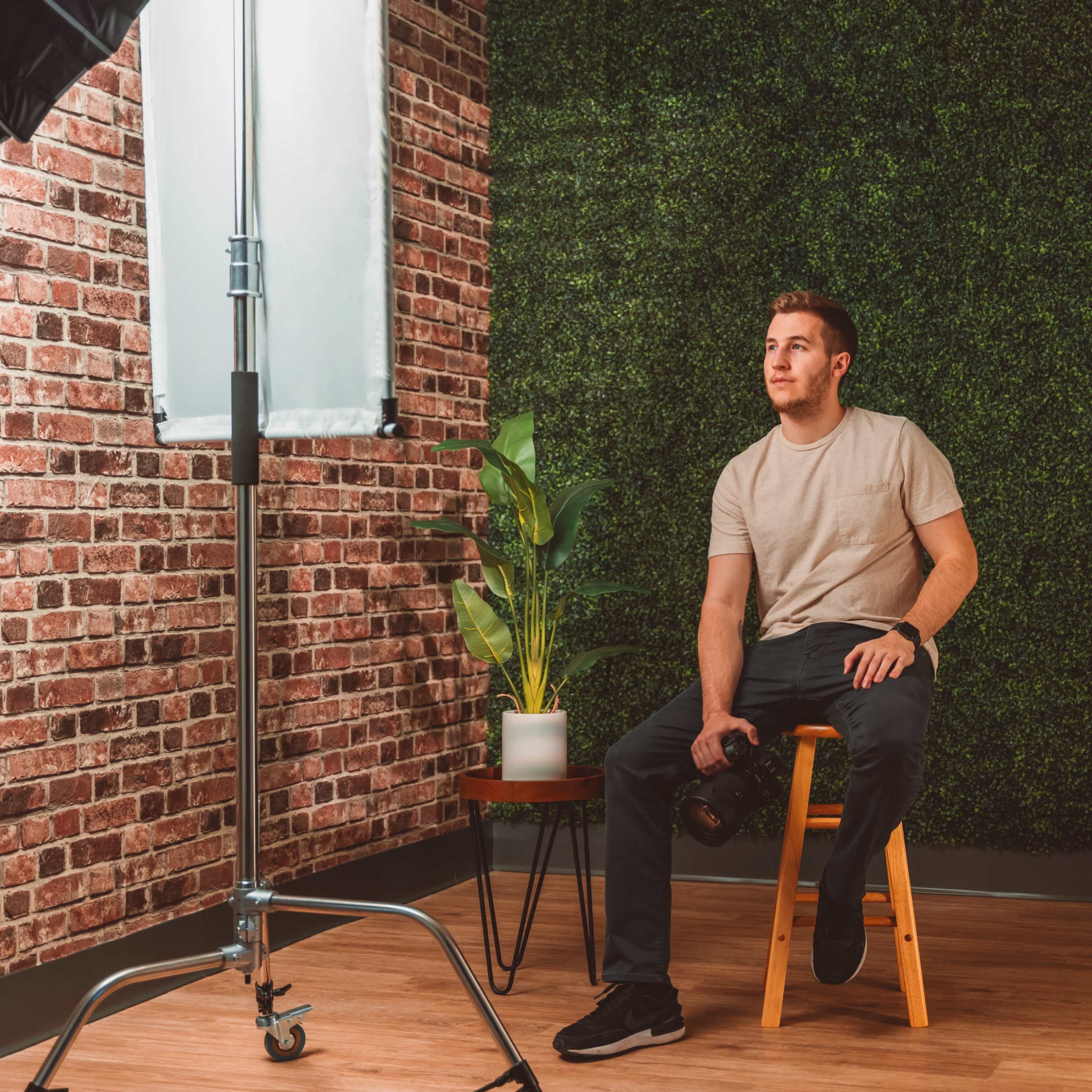 A man sits on a wooden stool holding a dumbbell in a studio with a backdrop featuring a brick wall and greenery.