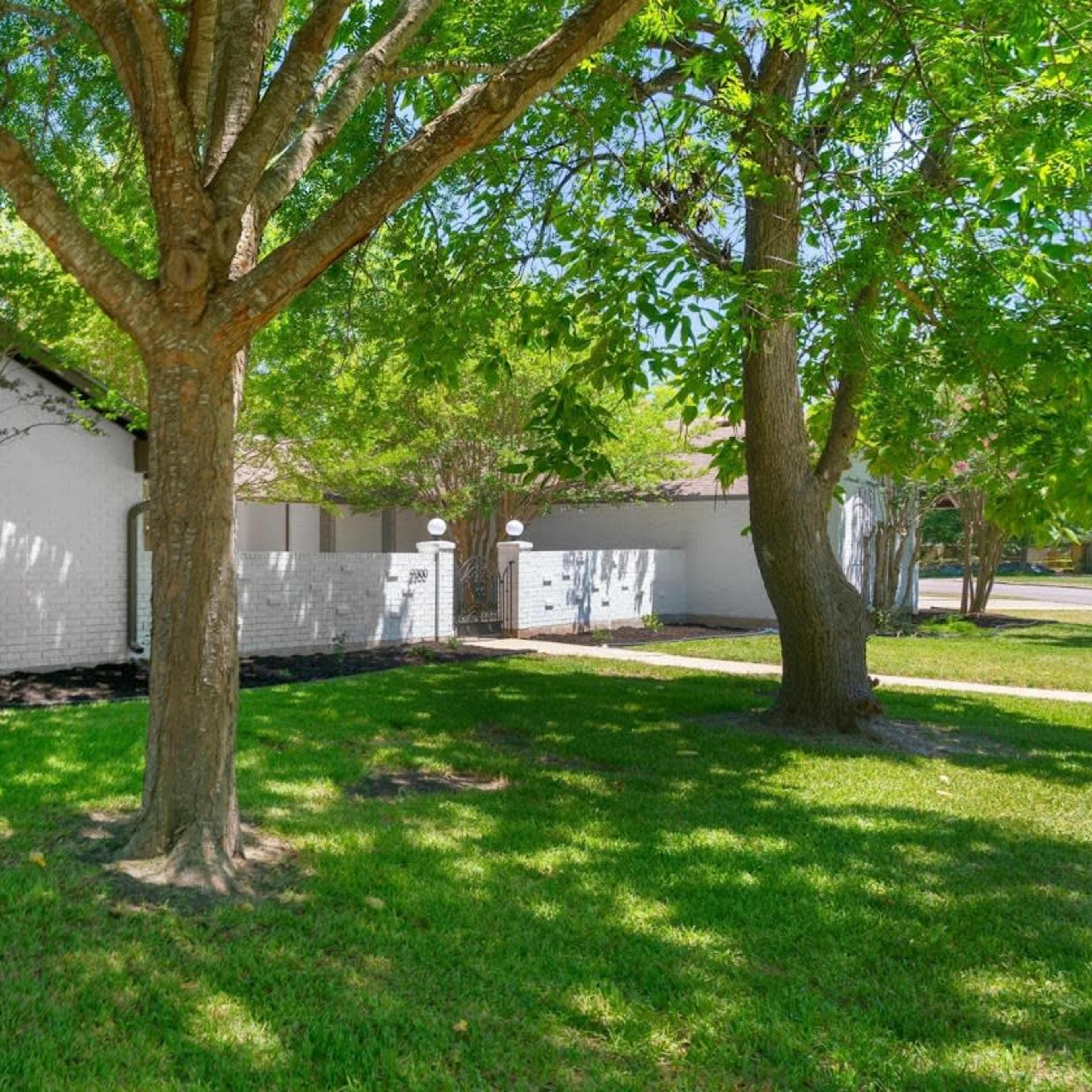 A white house with a sloped roof is set behind a green lawn and a tree, surrounded by a fence.