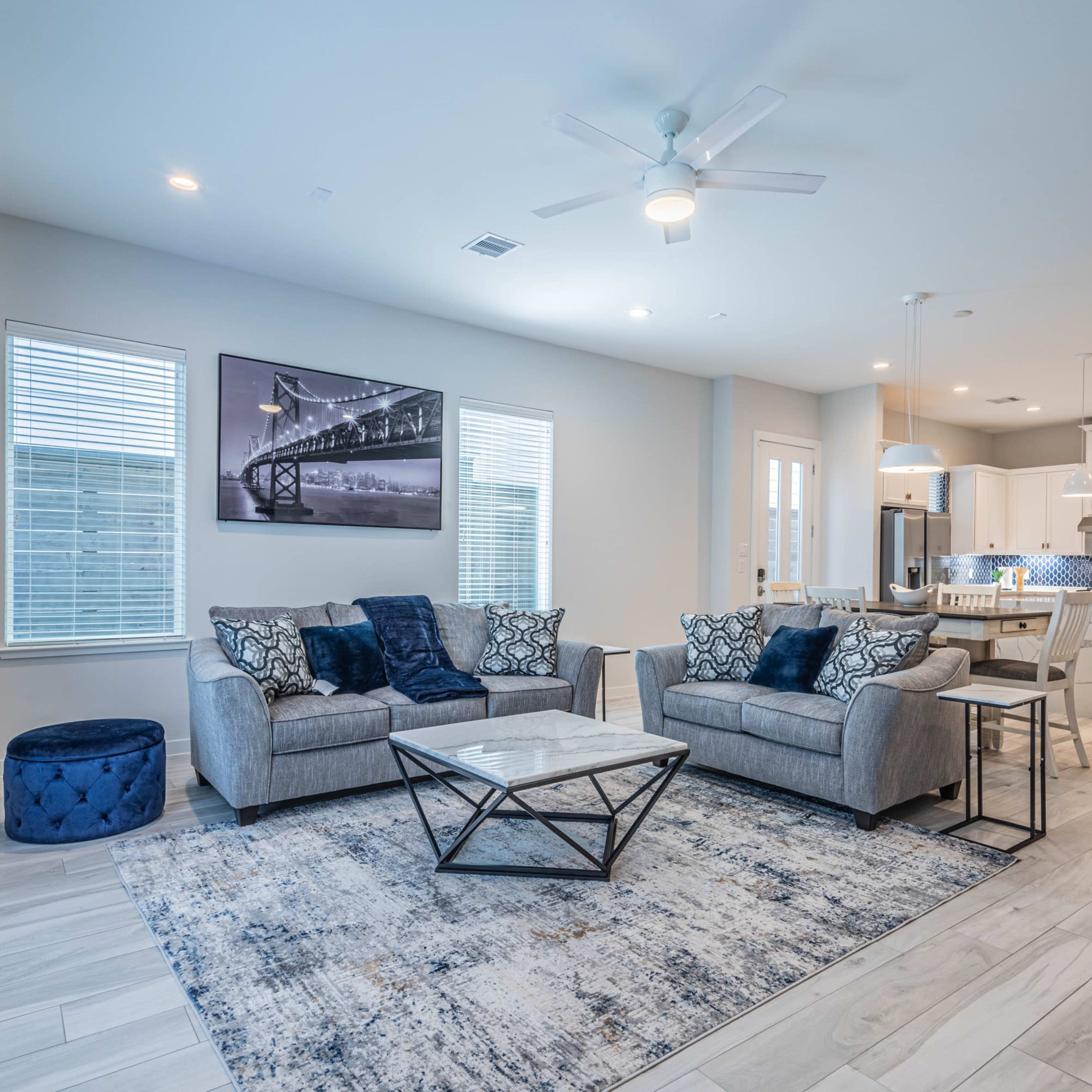 A modern living room with gray sofas, a coffee table, and a dining area visible in the background, all framed by blue decor and well-lit windows.