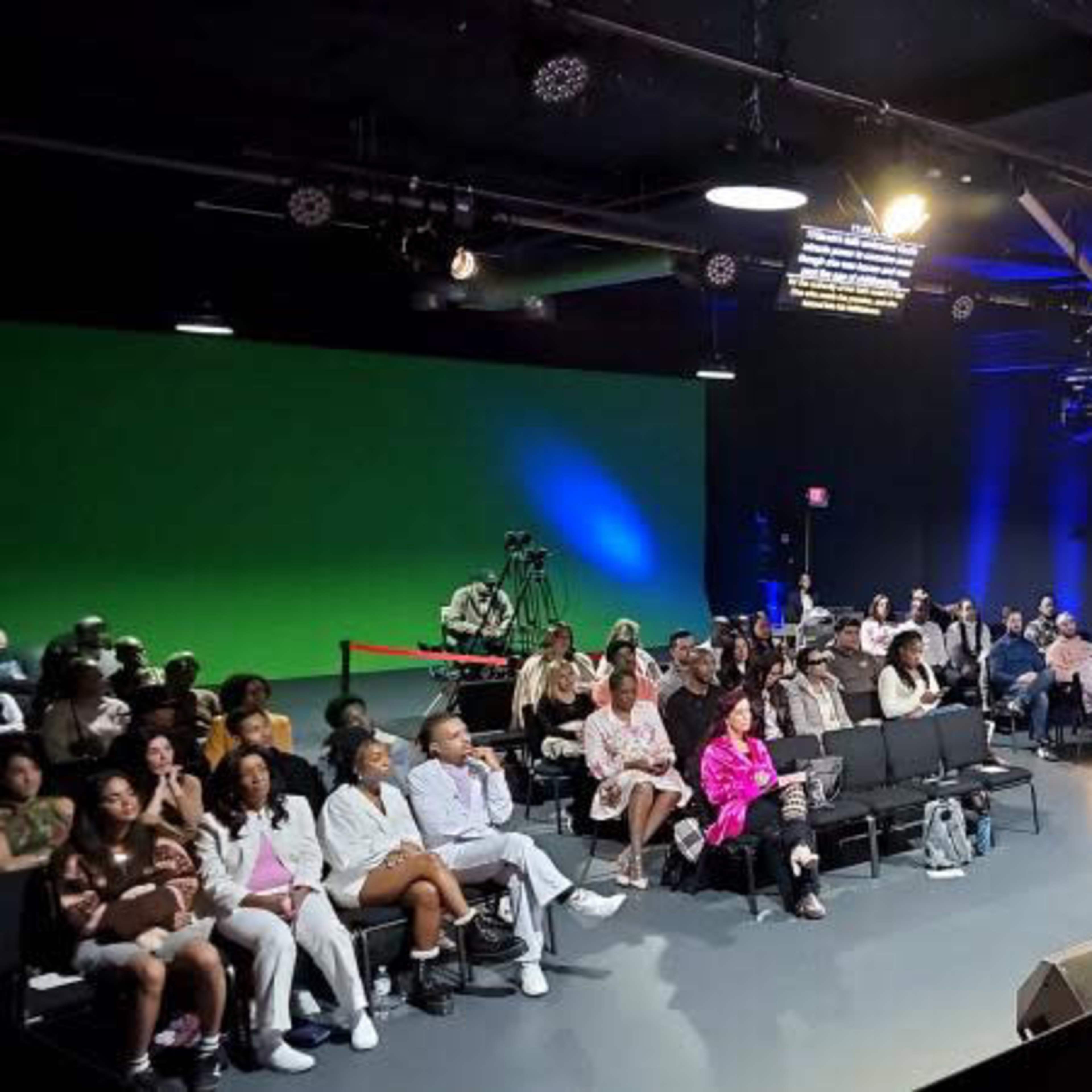A speaker is addressing an audience in a studio setting with green screens, projectors, and lighting.