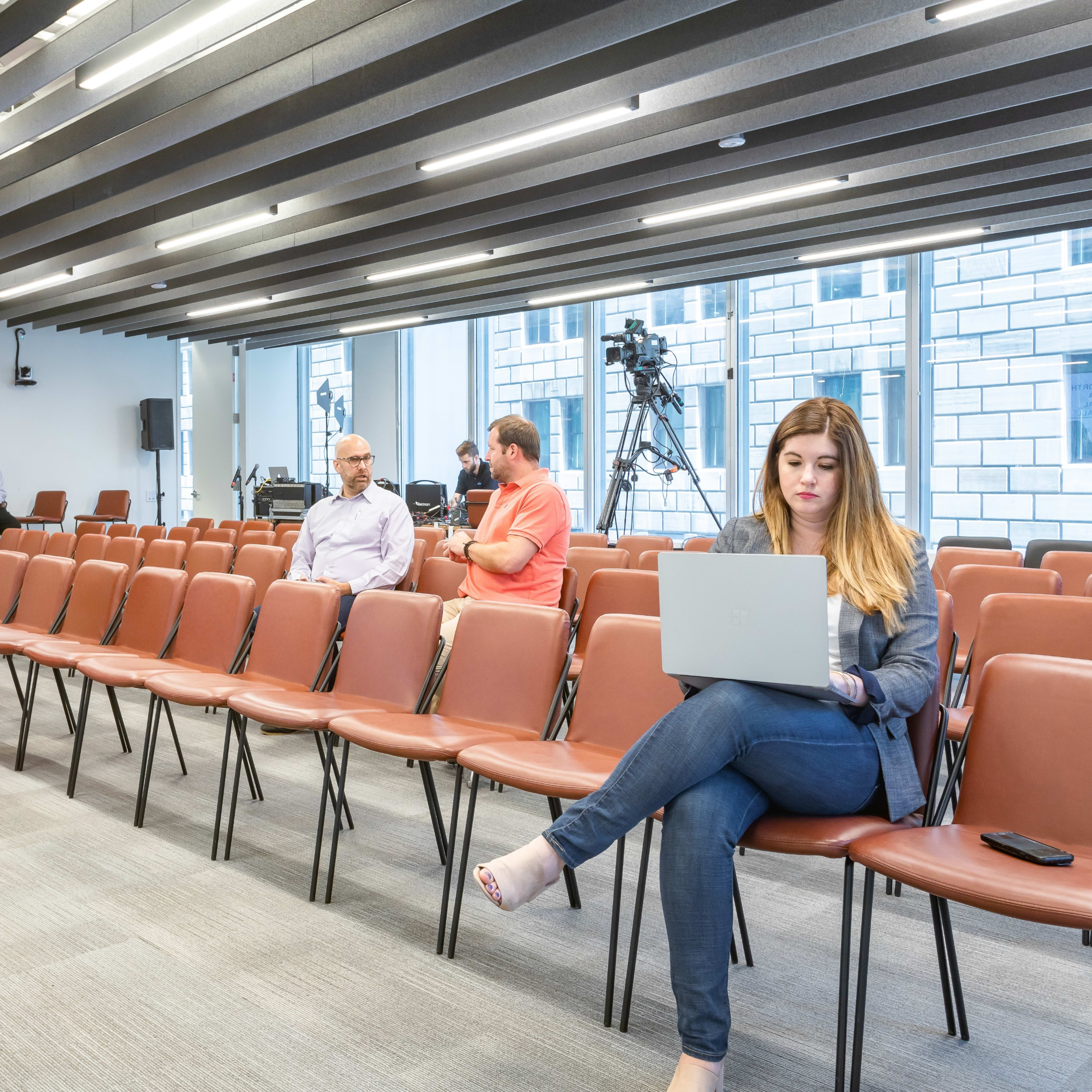 A largely empty conference room with rows of brown chairs, where a woman is sitting with a laptop and a few people are conversing nearby.