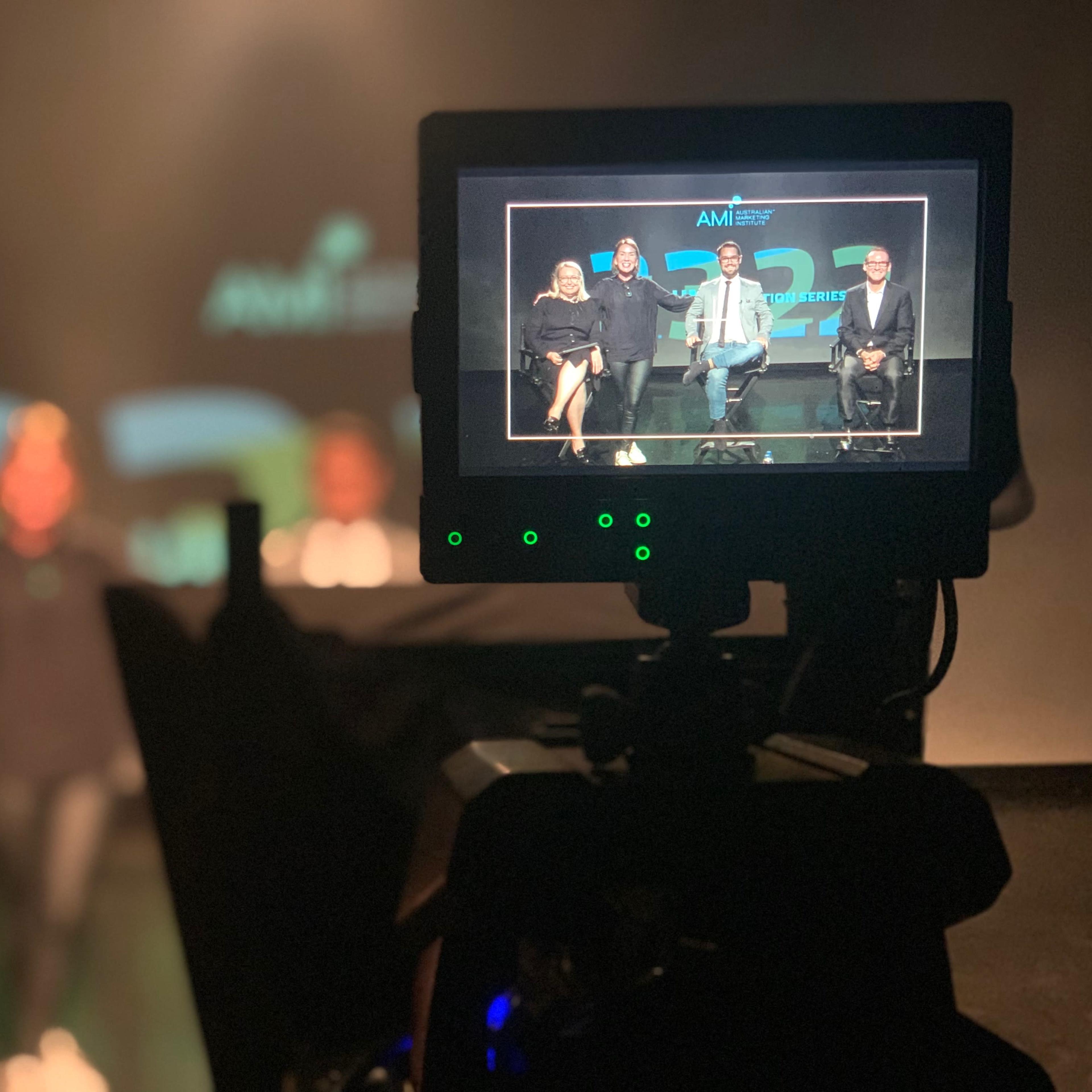 A camera captures a group of four people seated on a stage during a presentation, with a screen displaying branded graphics in the background.