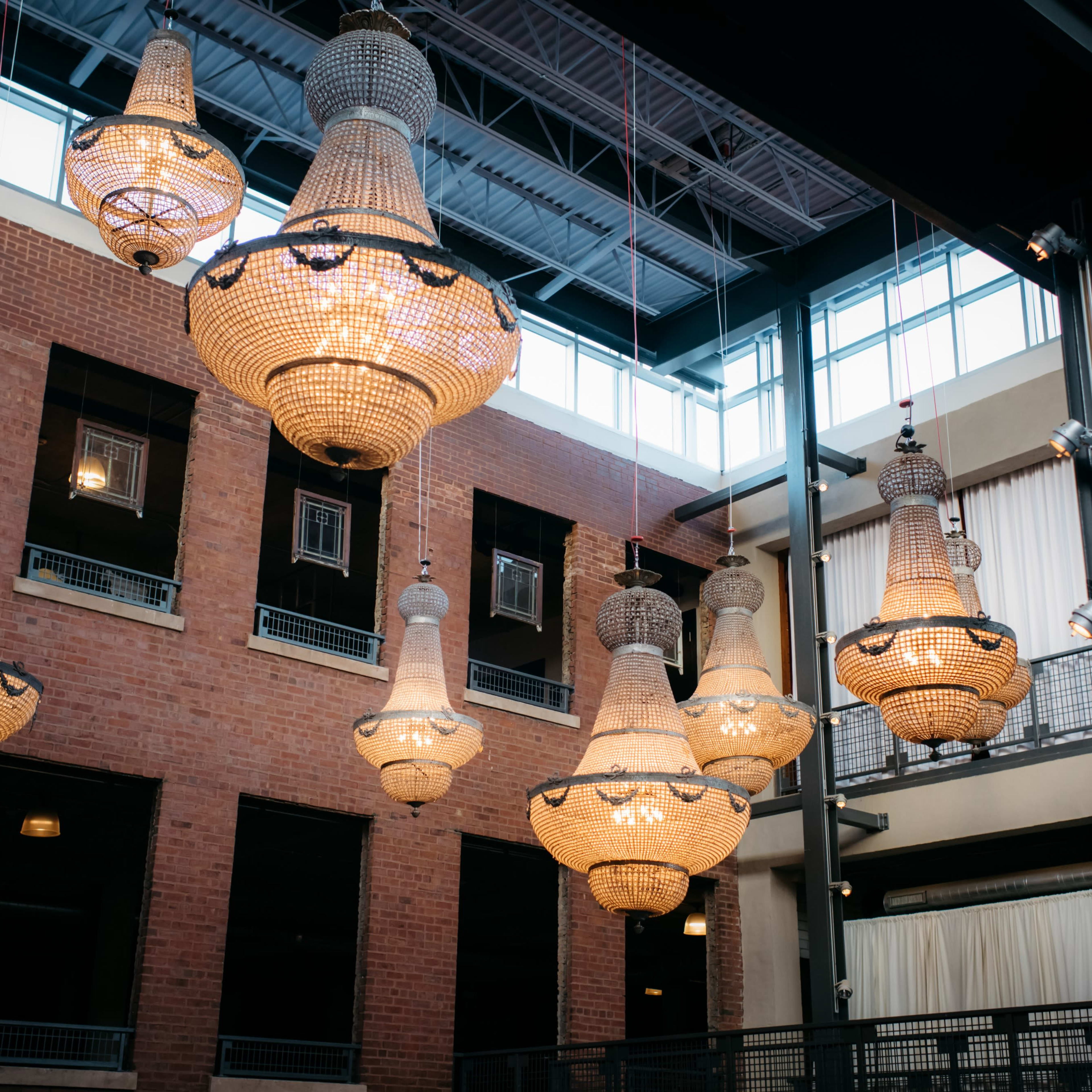 Multiple large, ornate chandeliers hanging from the ceiling of a building with brick walls and large windows.