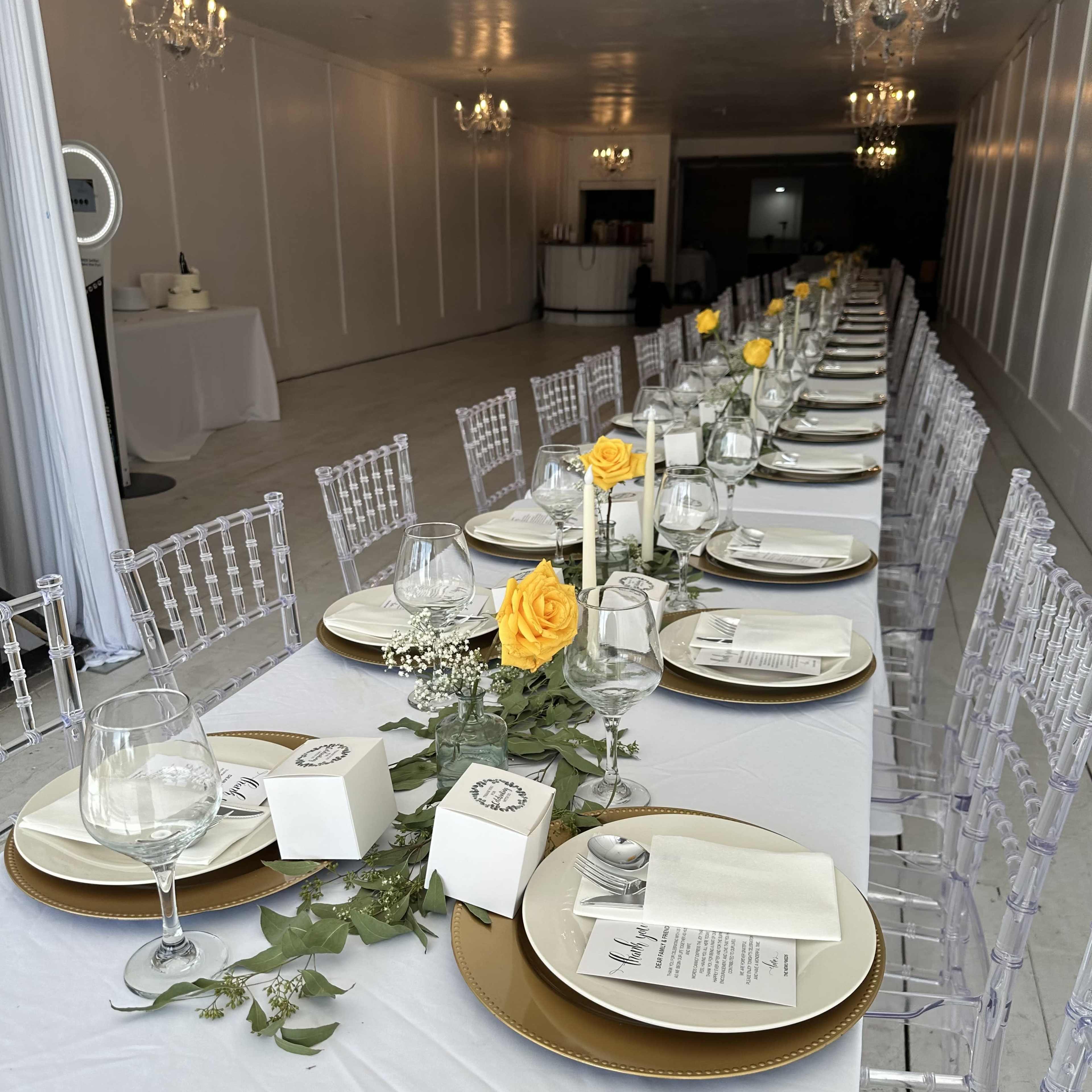 A long banquet table is elegantly set with yellow roses, glassware, and decorative items, arranged under chandeliers in a spacious room.