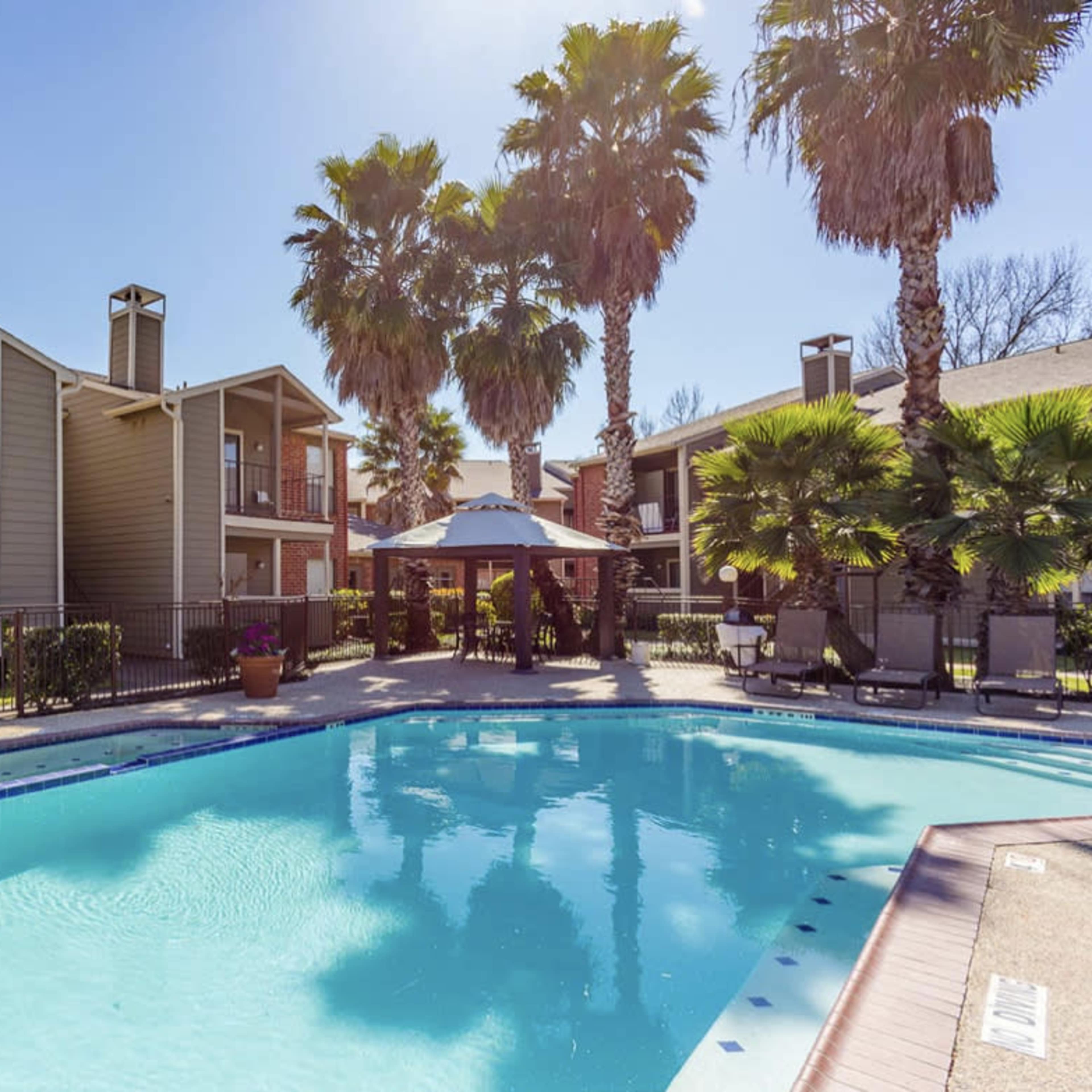 A swimming pool surrounded by palm trees and two-story apartment buildings.