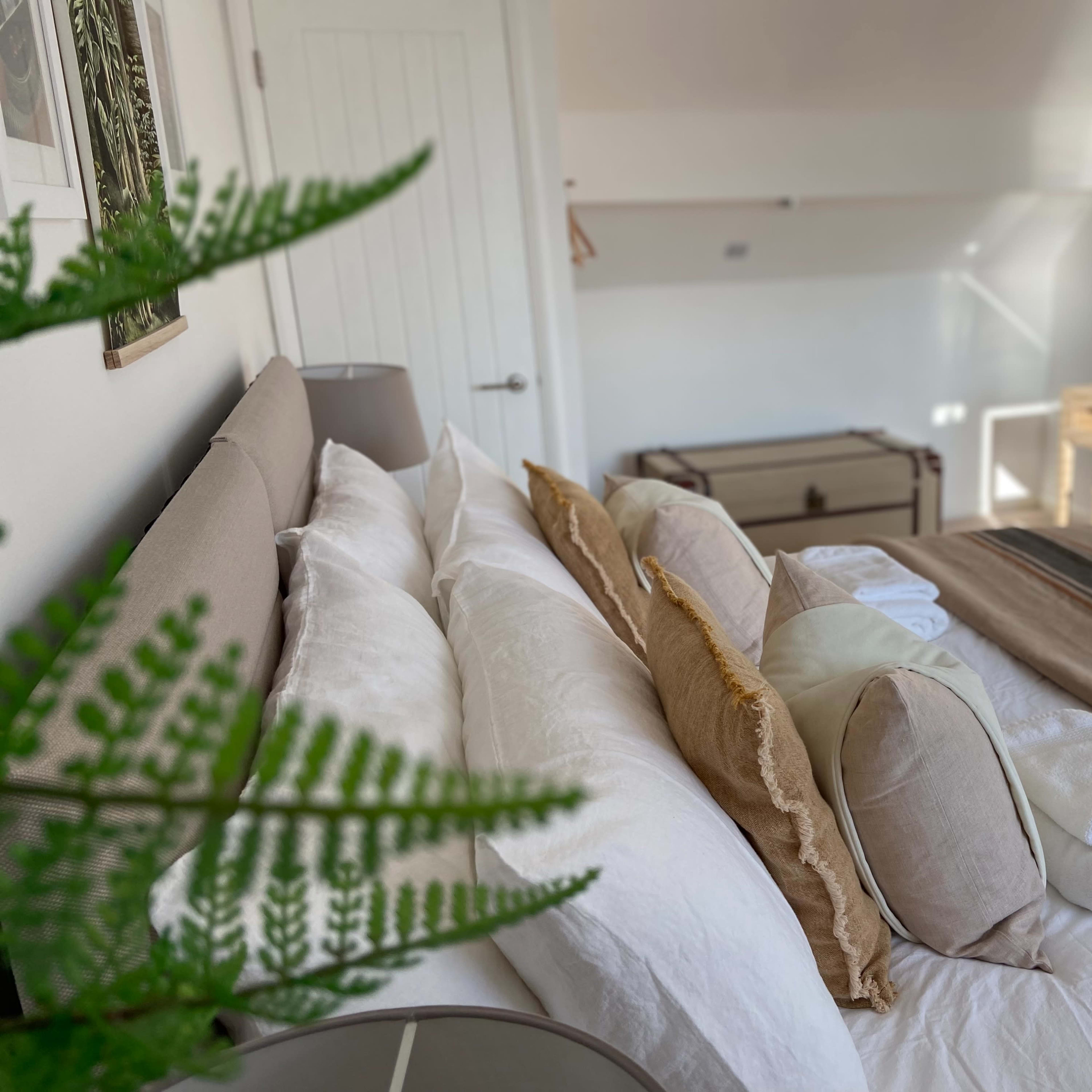 A neatly arranged bedroom features a bed with multiple pillows, a small dresser, and a trunk in the background.