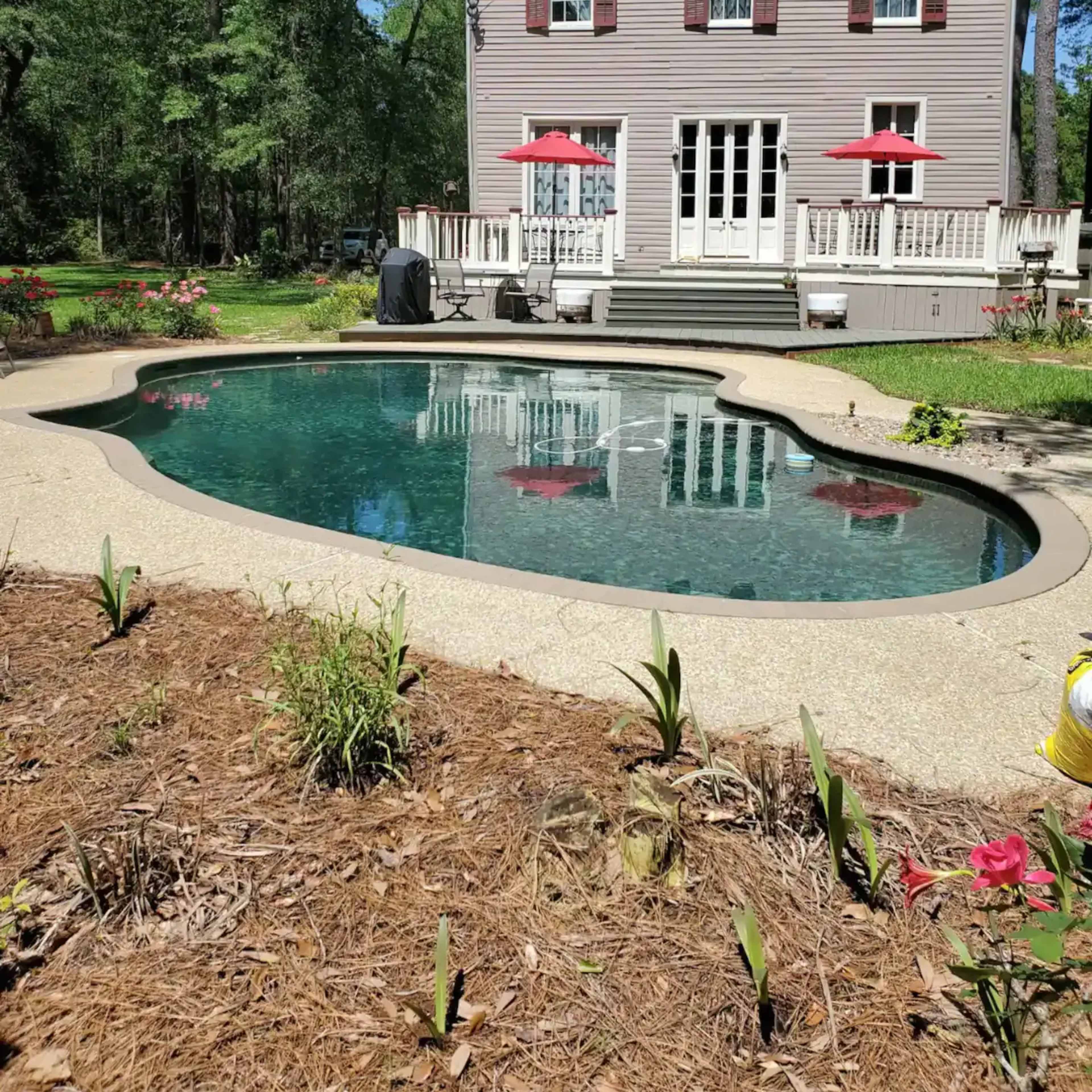 A large, two-story gray house with red shutters overlooks a curved swimming pool surrounded by landscaped gardens.