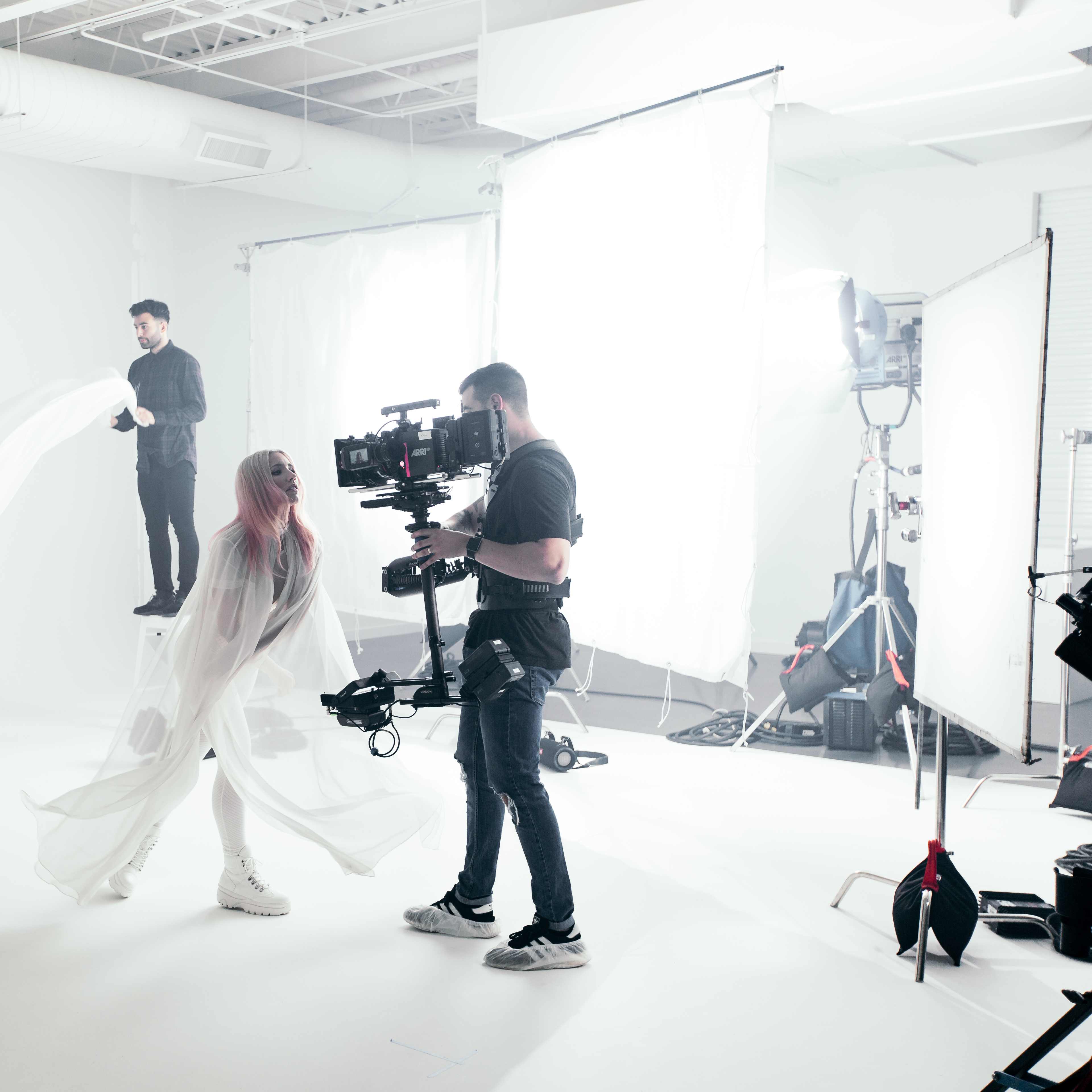 A film crew is capturing a scene in a brightly lit studio, featuring models draped in white fabric and a cameraman filming with a stabilizer.