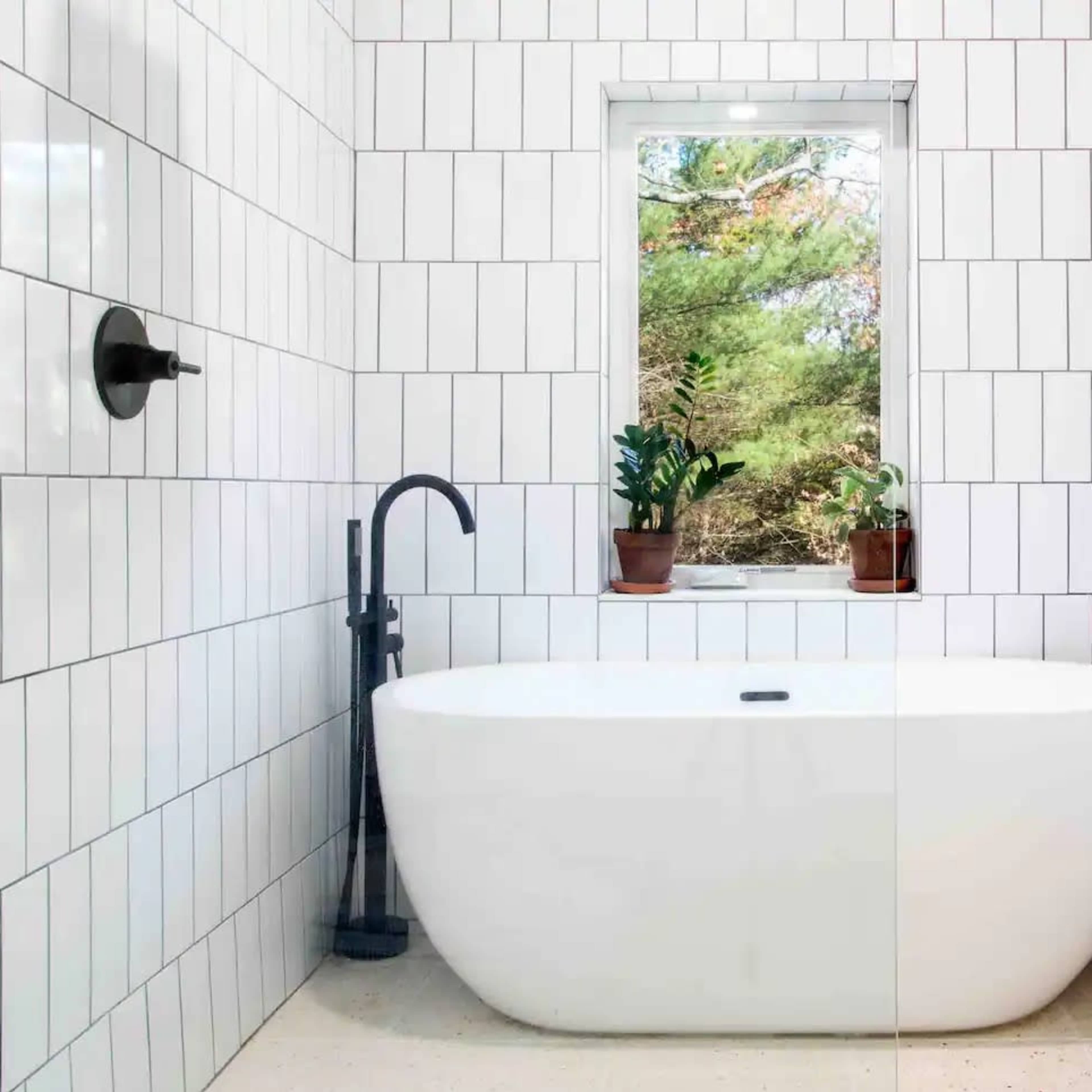 A modern bathroom features a freestanding white bathtub surrounded by white tiled walls and a window with potted plants.