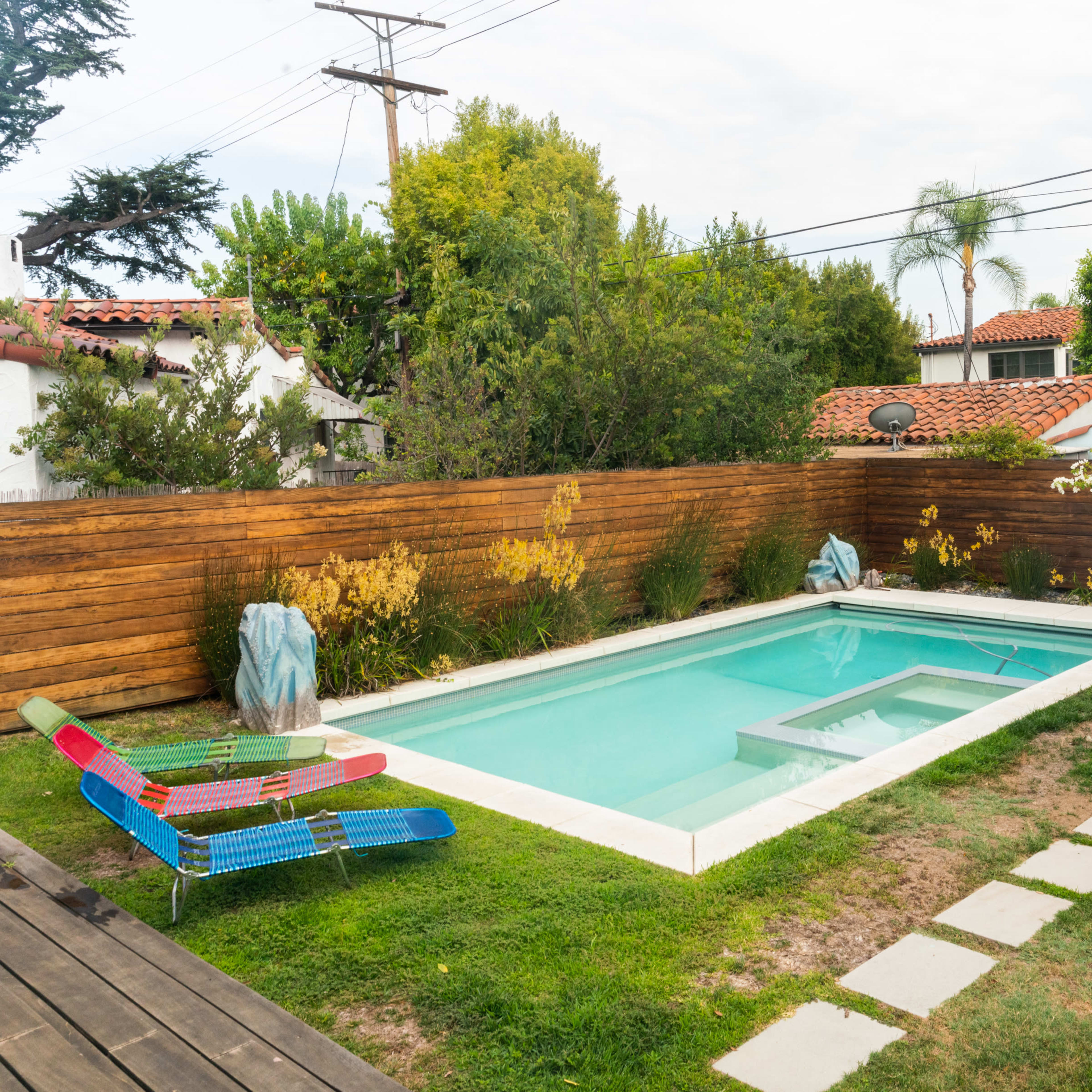 A shallow rectangular pool surrounded by grass and wooden fencing, with a colorful lounge chair and nearby greenery.