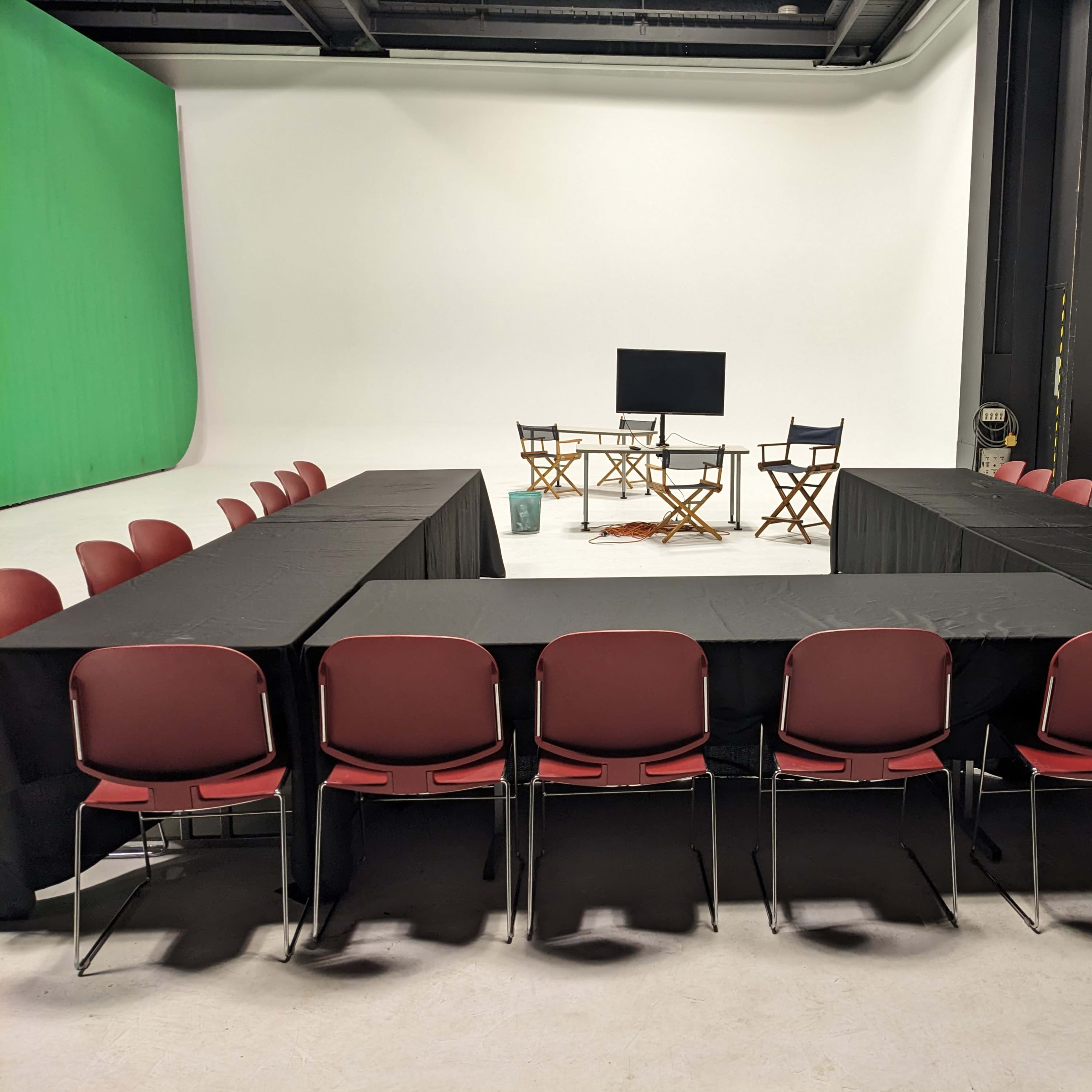 A studio setup features a rectangular arrangement of tables with chairs around them, a screen, and director's chairs at one end.