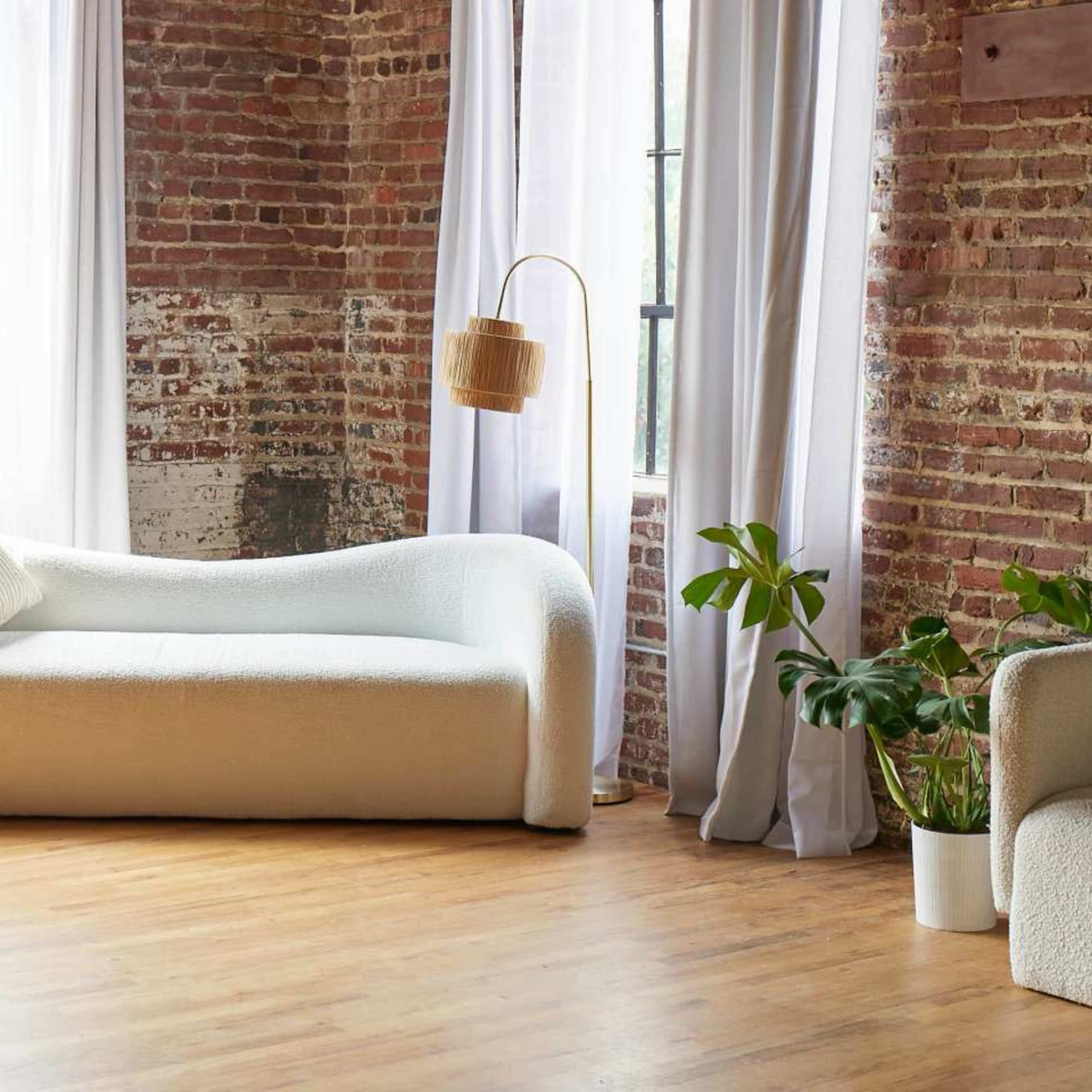 A curved white sofa is positioned in a room with exposed brick walls, large windows, a floor lamp, and a potted plant.
