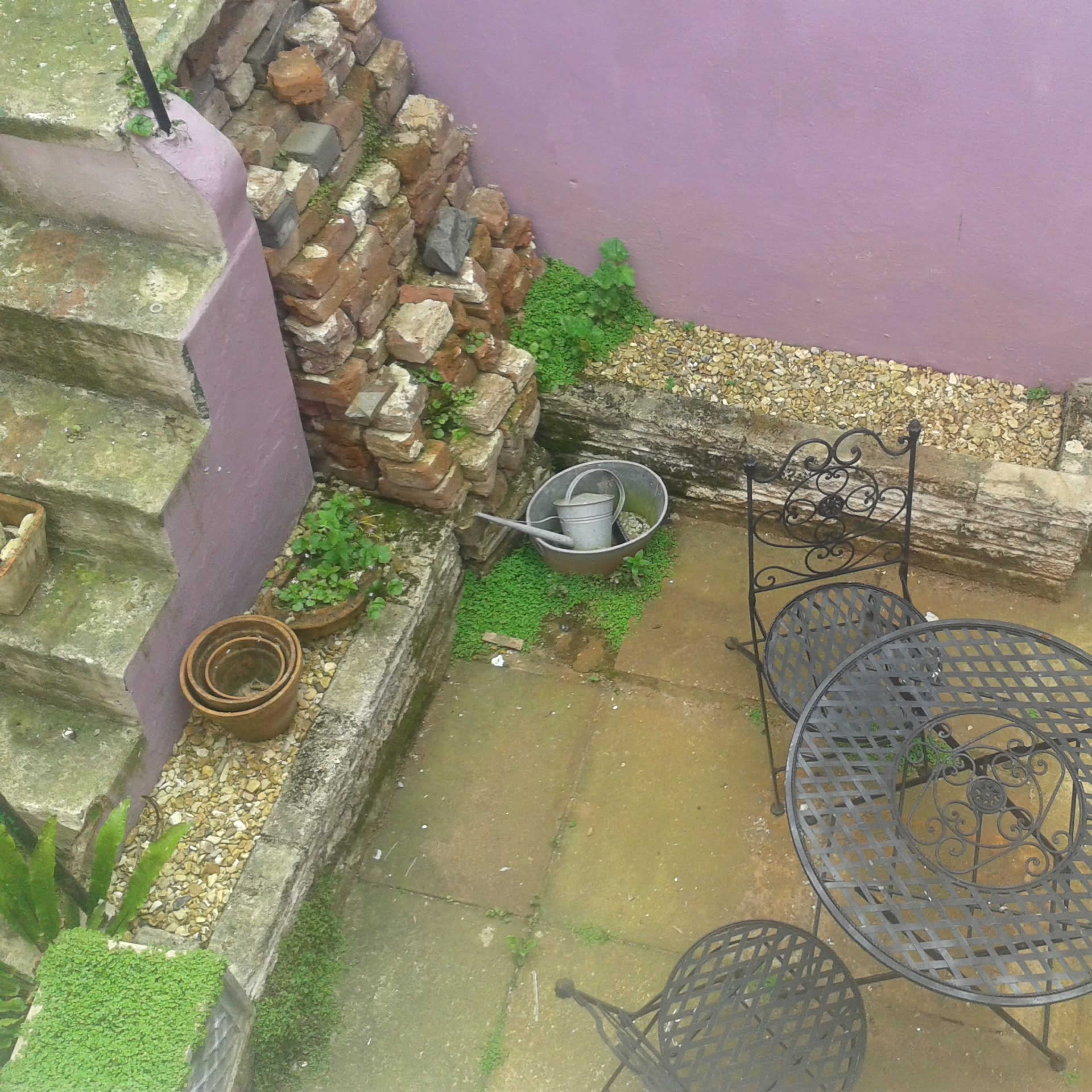 A small courtyard features a black metal table and chairs, surrounded by stone steps, a brick wall, and scattered pots.