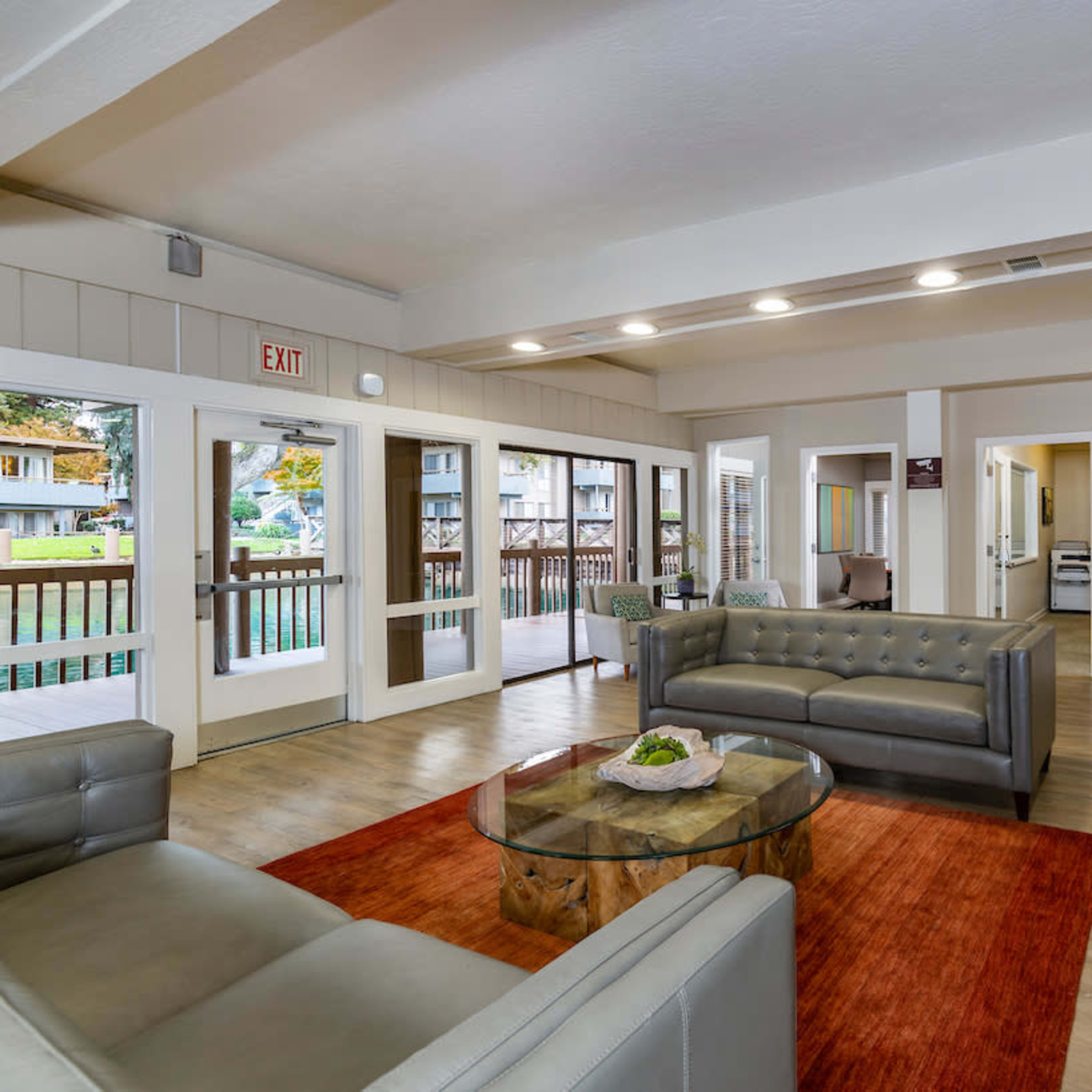 A modern living room featuring gray sofas, a glass coffee table, and large windows allowing natural light, with doors leading to a balcony area.