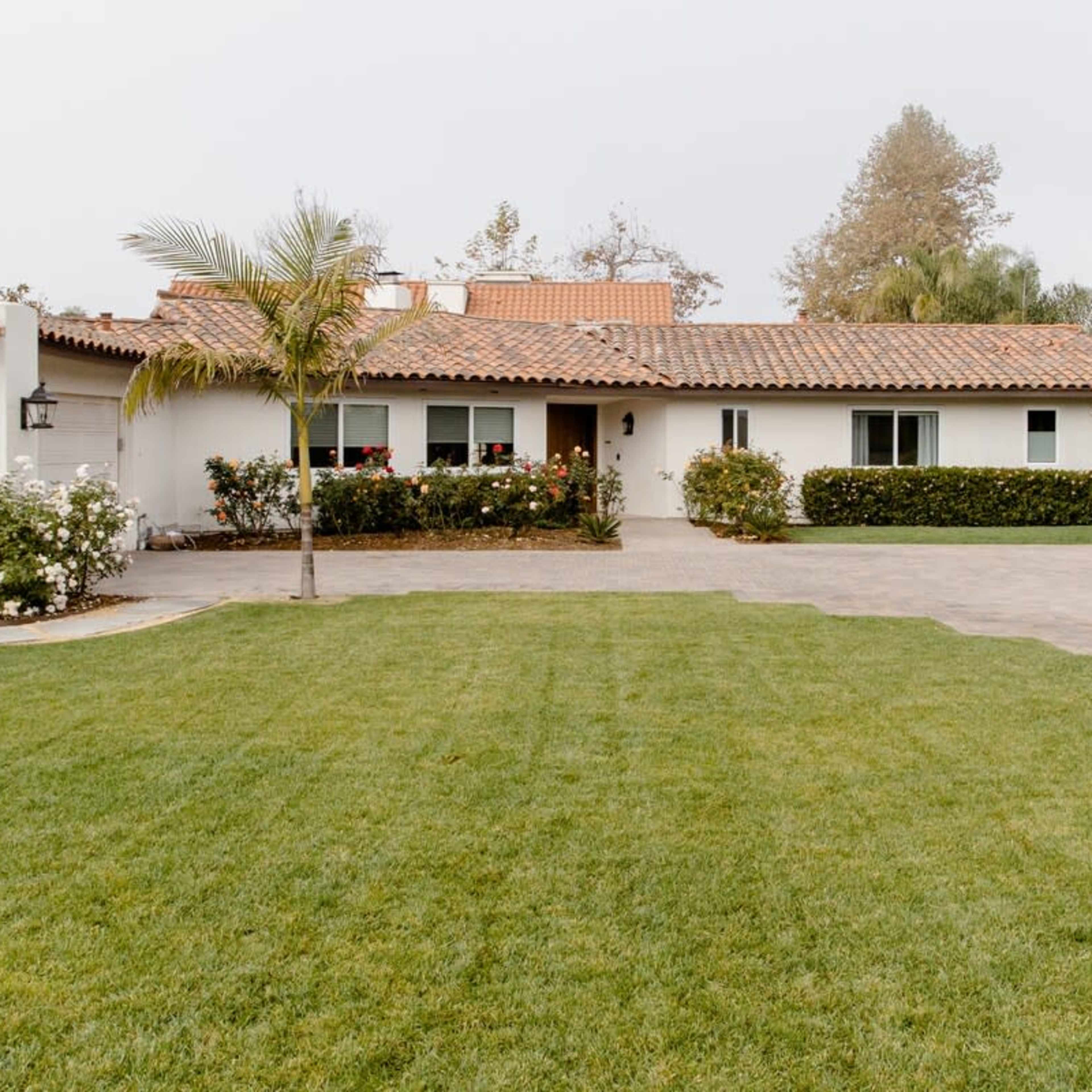 A one-story house with a tiled roof is set behind a manicured lawn and surrounded by shrubbery and flowers.