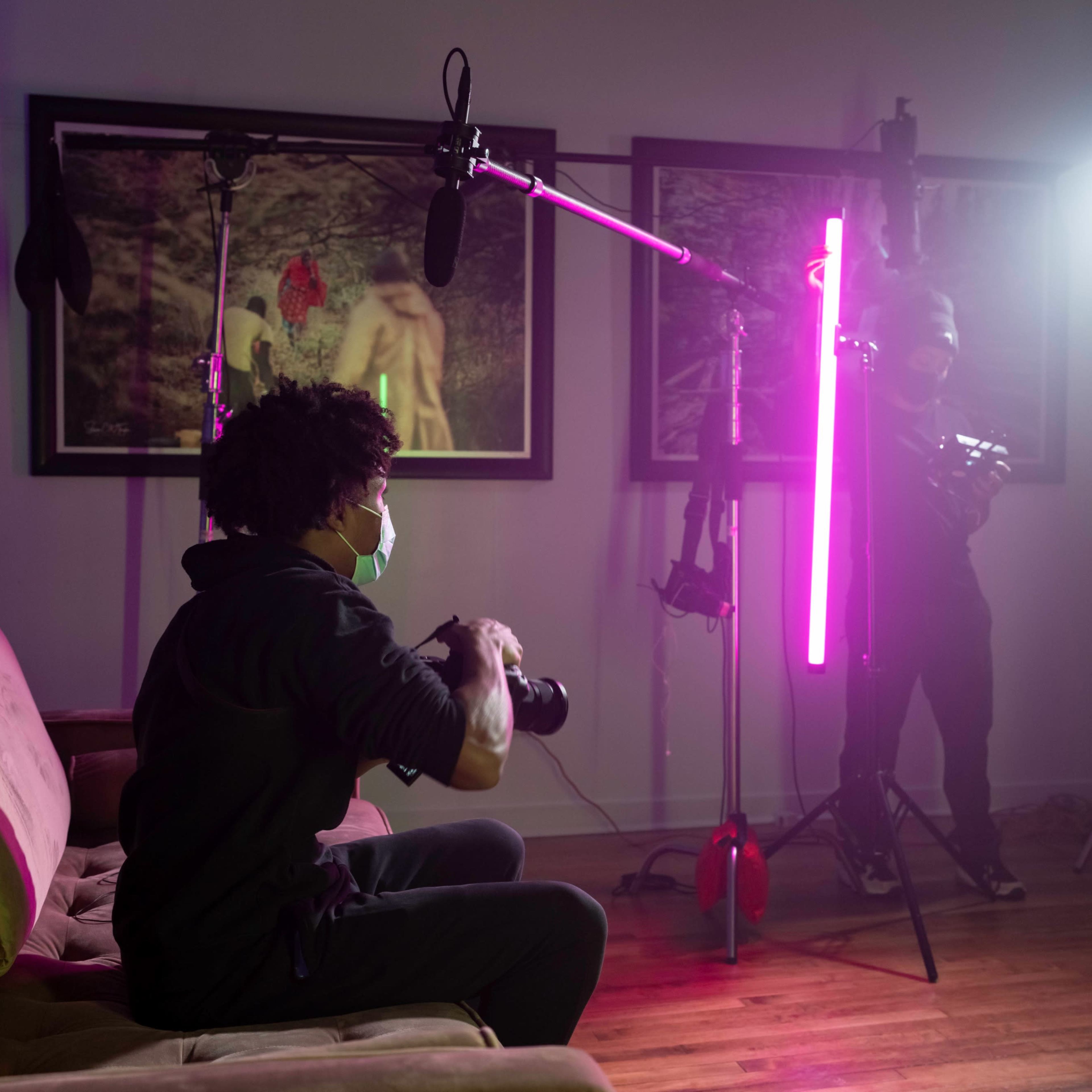 A photographer sits on a couch with a camera, while crew members set up lighting and equipment in a studio.