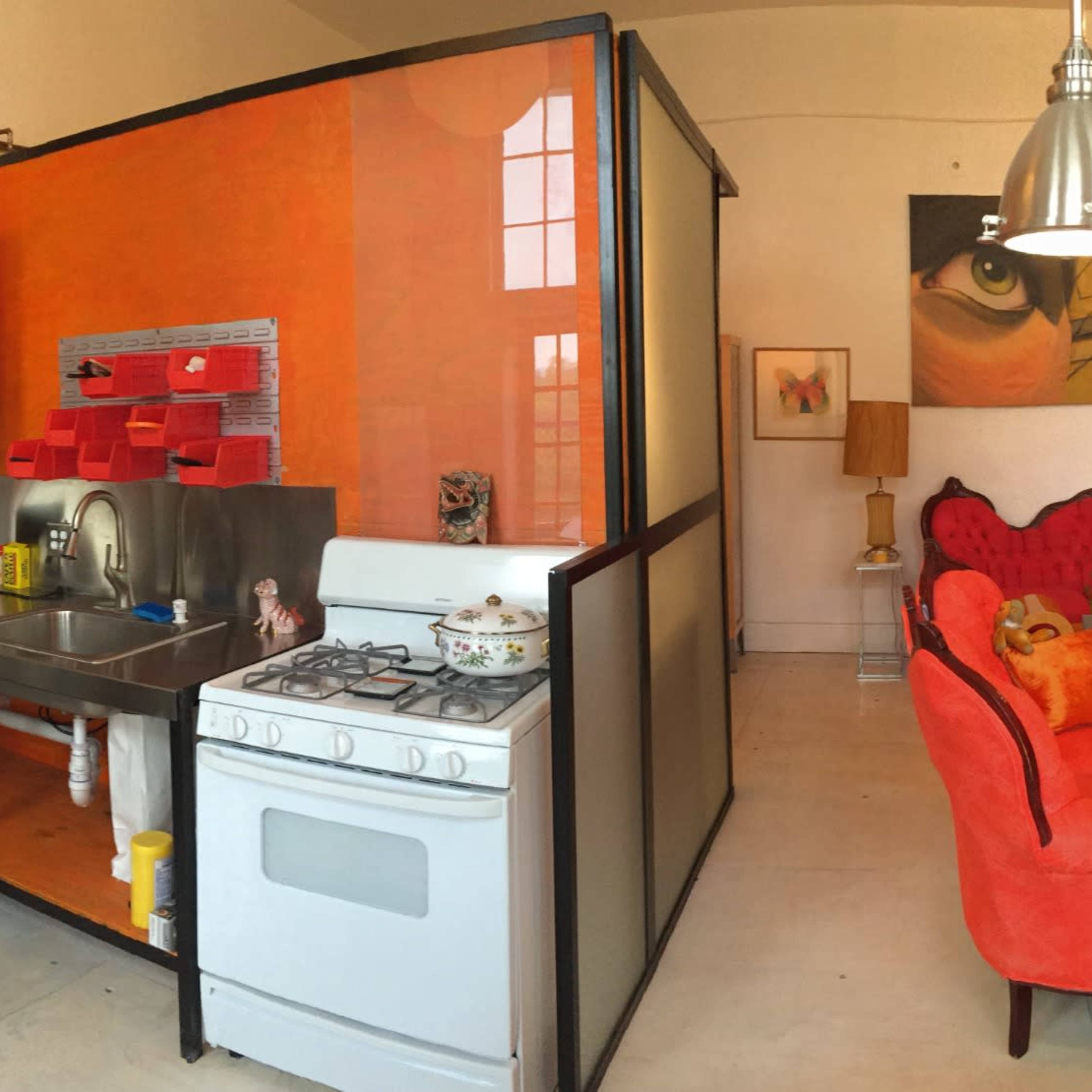 A panoramic view of a stylish living space featuring a kitchen with modern appliances, a vibrant orange accent wall, a cozy seating area with red furniture, and a decorative wooden cabinet.
