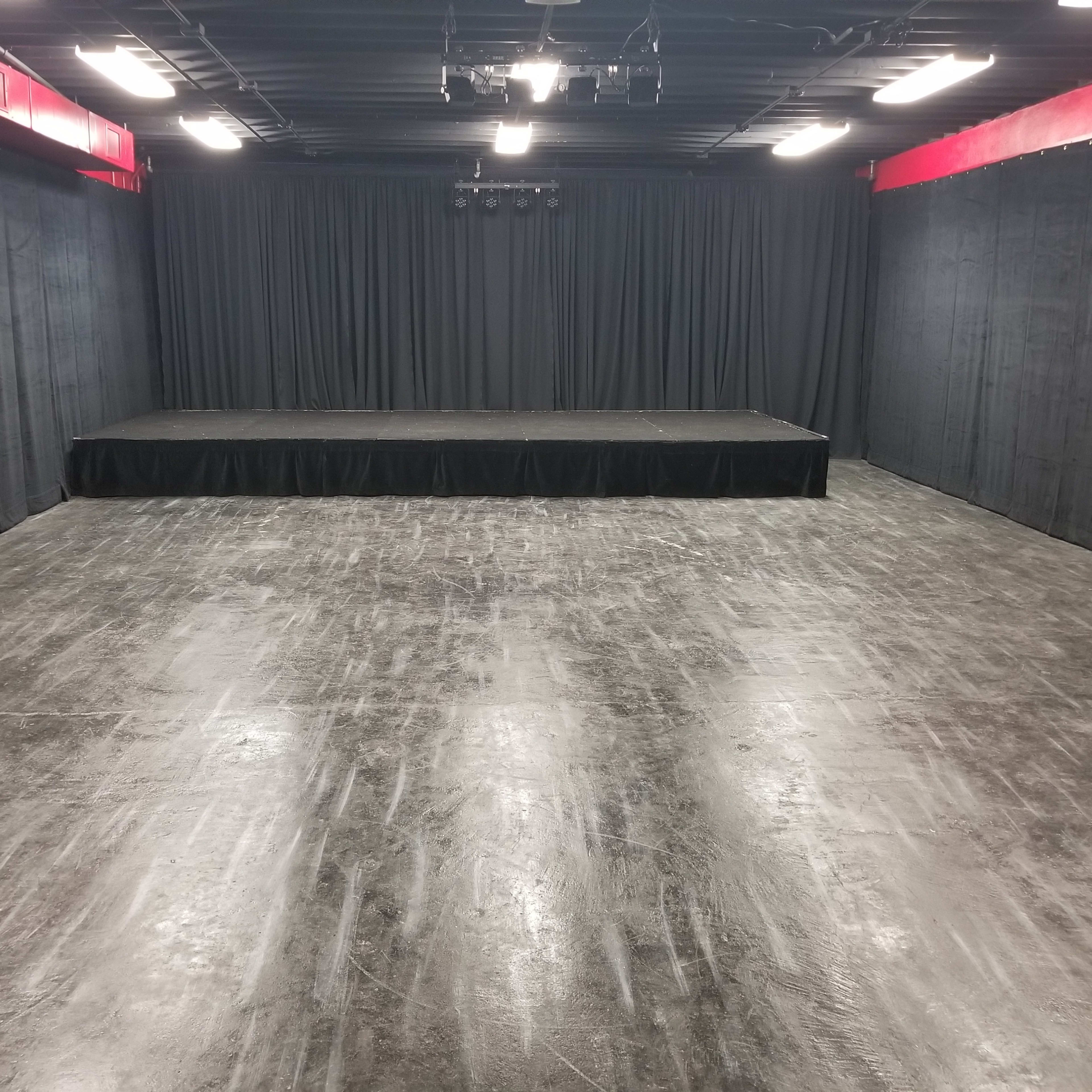 A dark, empty stage area with black curtains, a raised platform in the center, and a polished concrete floor.