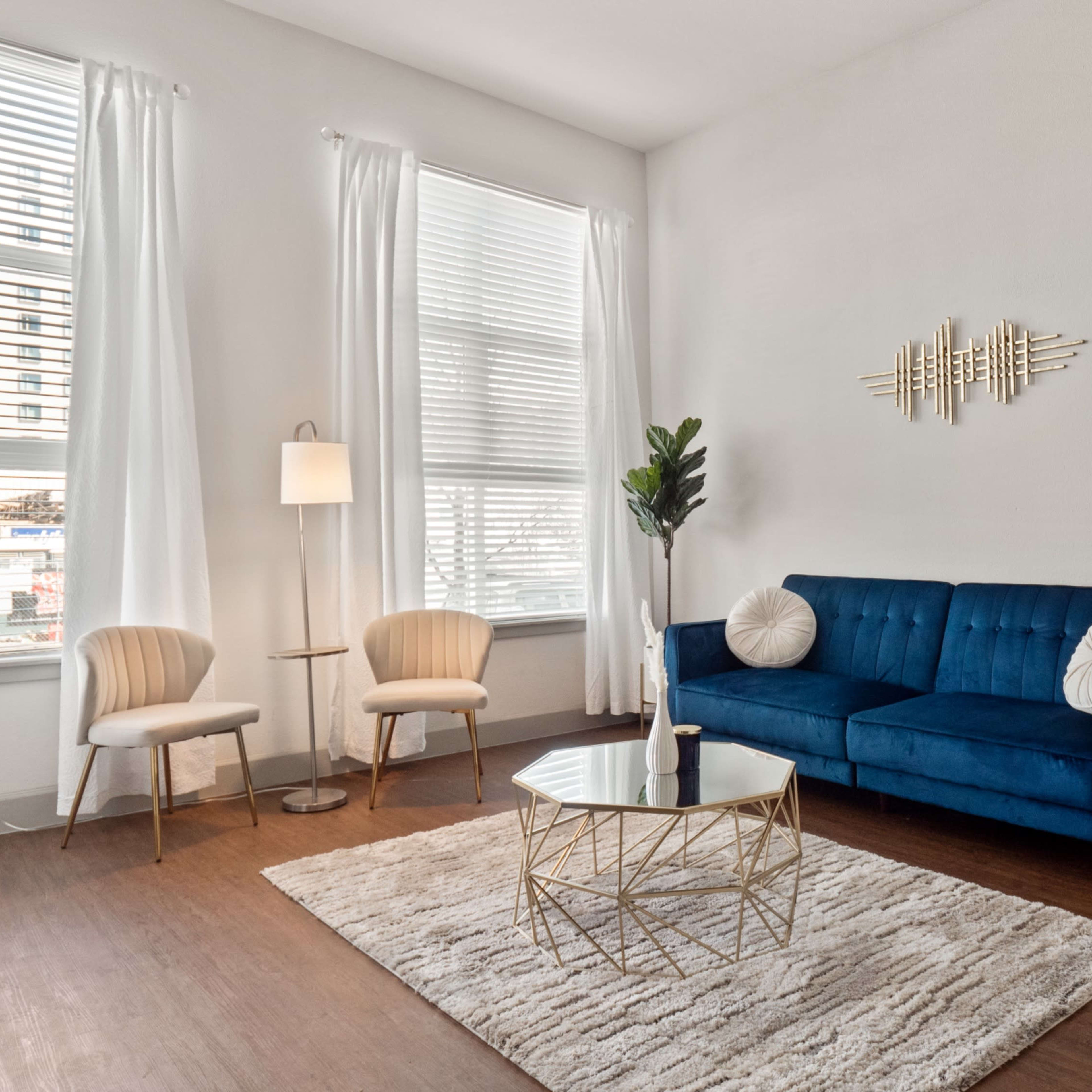 A bright living room featuring a blue couch, a round coffee table, two light-colored chairs, and large windows with white curtains.