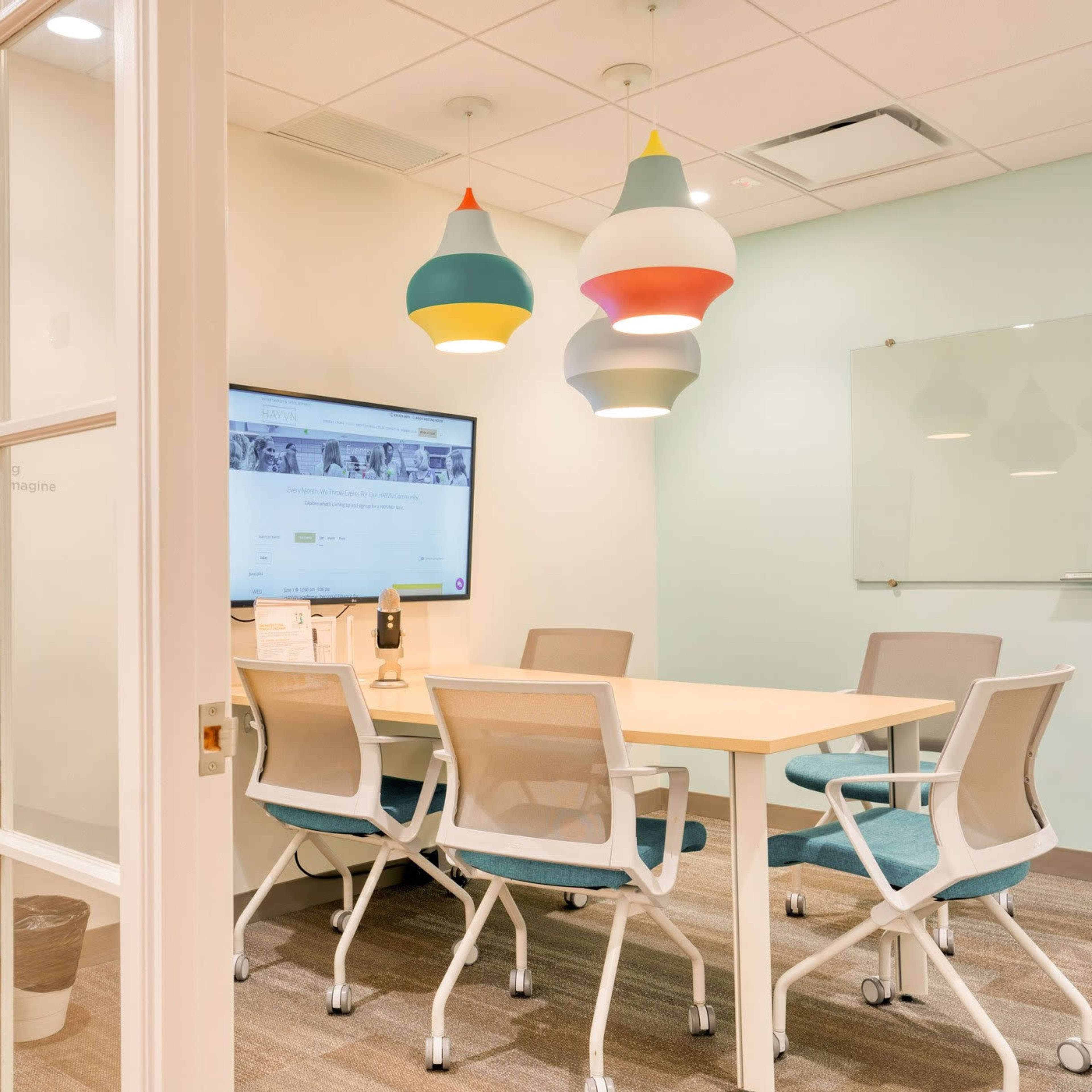 A small conference room features a central table surrounded by four chairs, a large screen on one wall, and colorful pendant lights overhead.