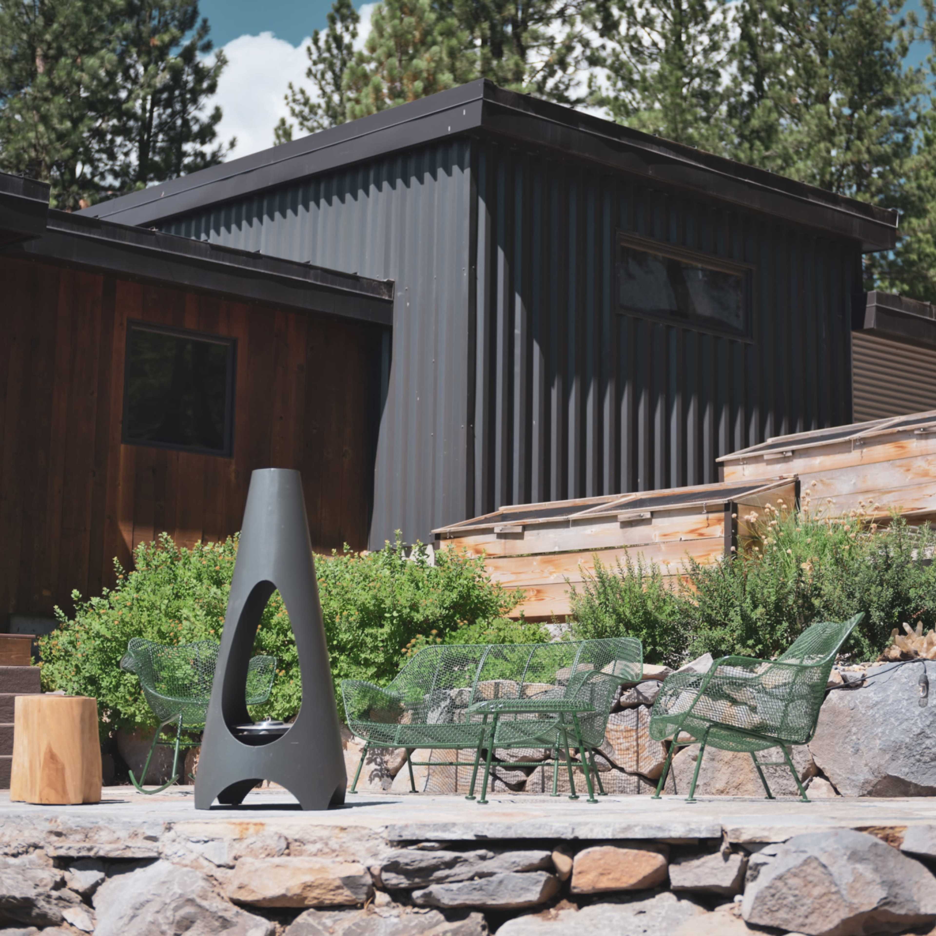 A modern outdoor area with a charcoal-colored fire pit, green chairs, and wooden planters, set against a backdrop of two contemporary buildings and trees.
