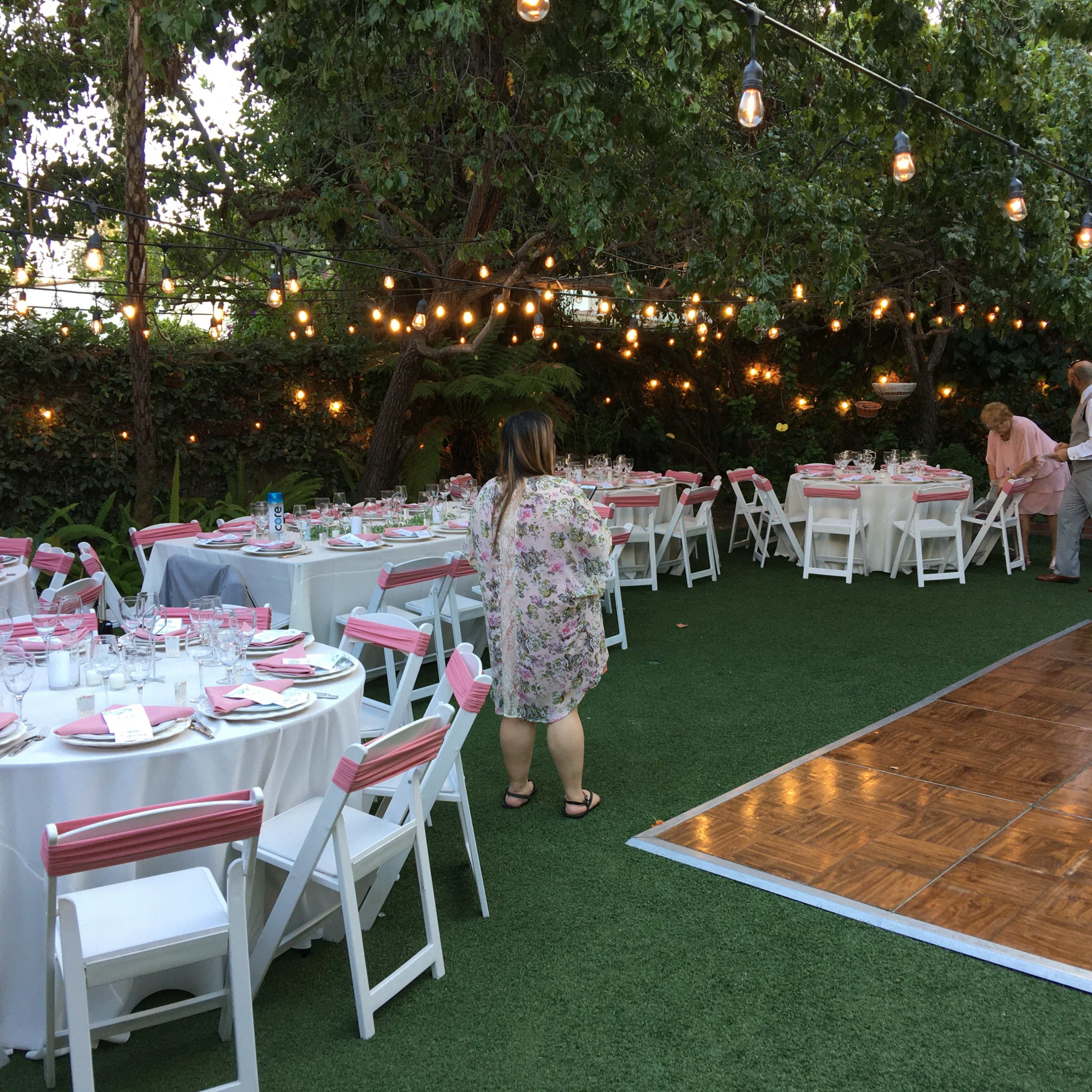 A garden setting features tables set with pink and white decorations beneath string lights, with a dance floor to the side.