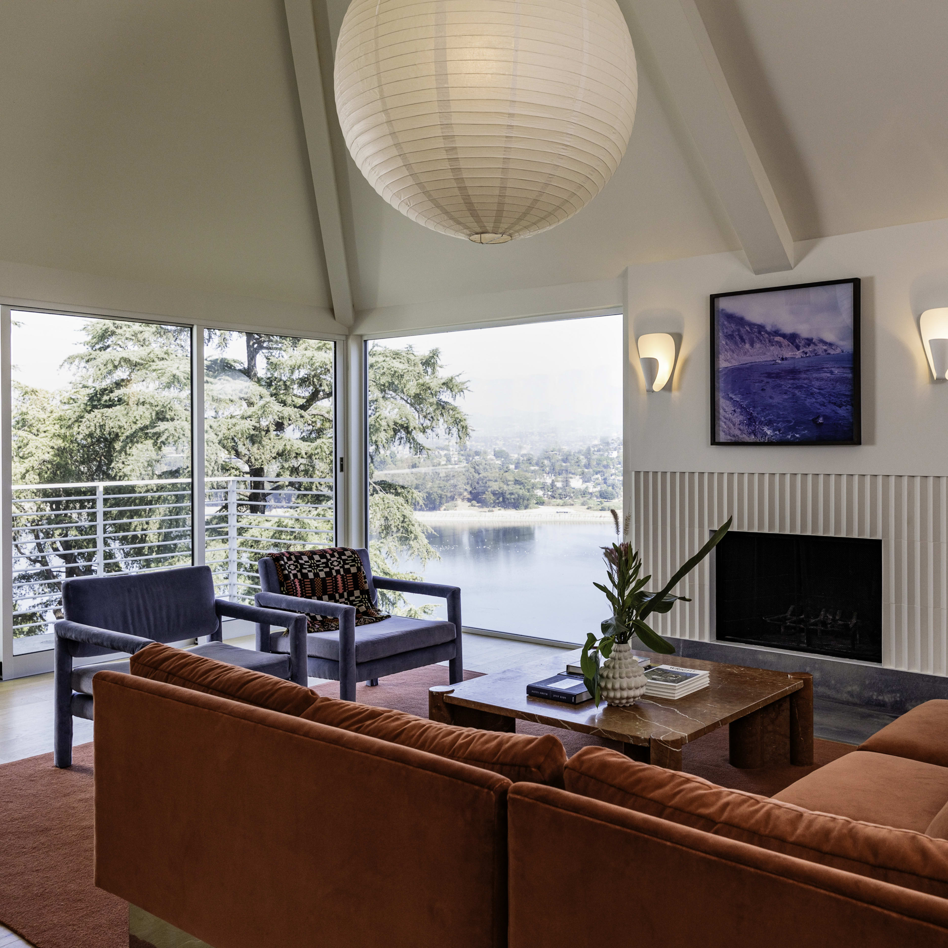 A modern living room features a large, orange sofa, two chairs, and a coffee table, all with a view of a body of water through expansive windows.