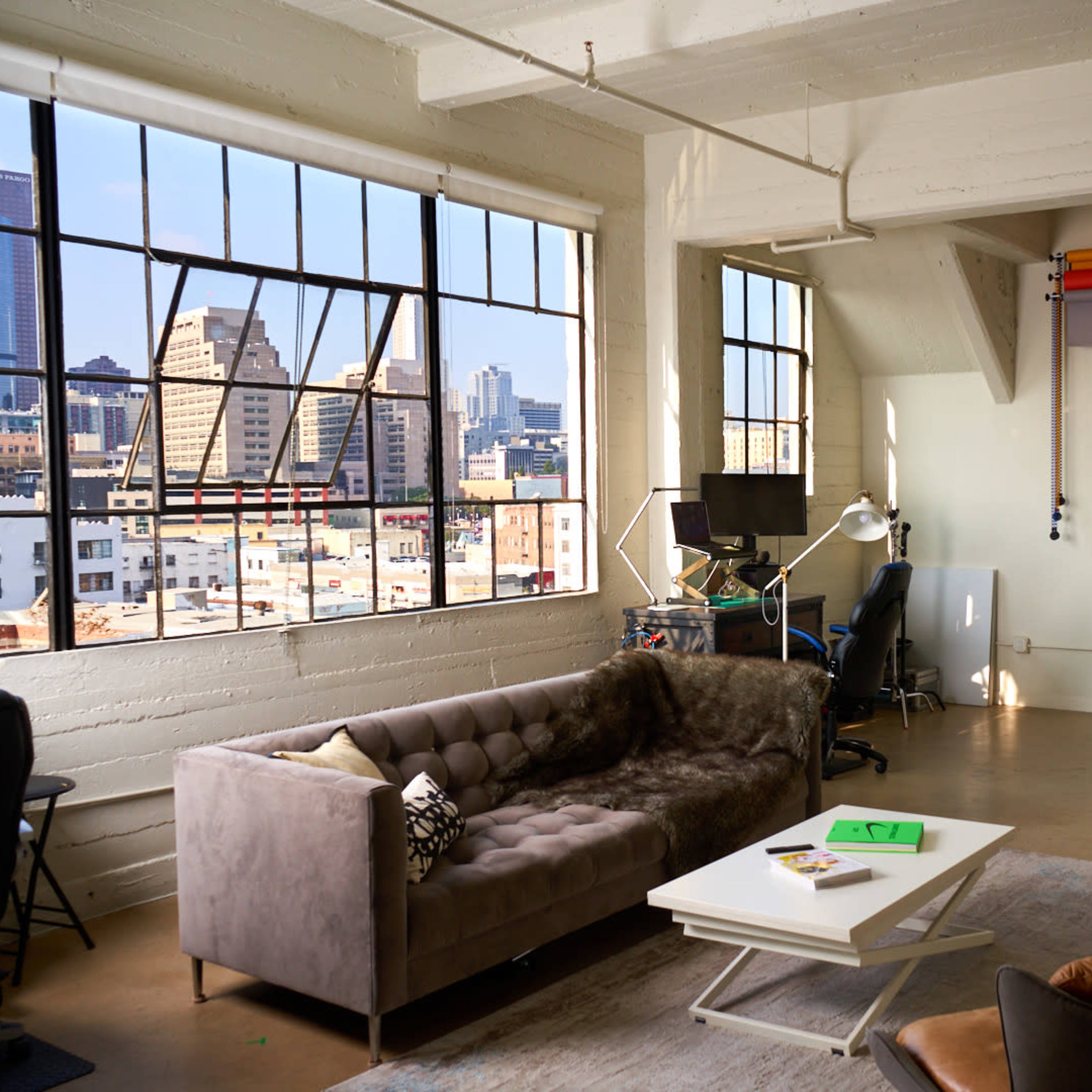 A modern, spacious loft-style living area with large windows, a gray couch, and a coffee table, overlooking a city skyline.