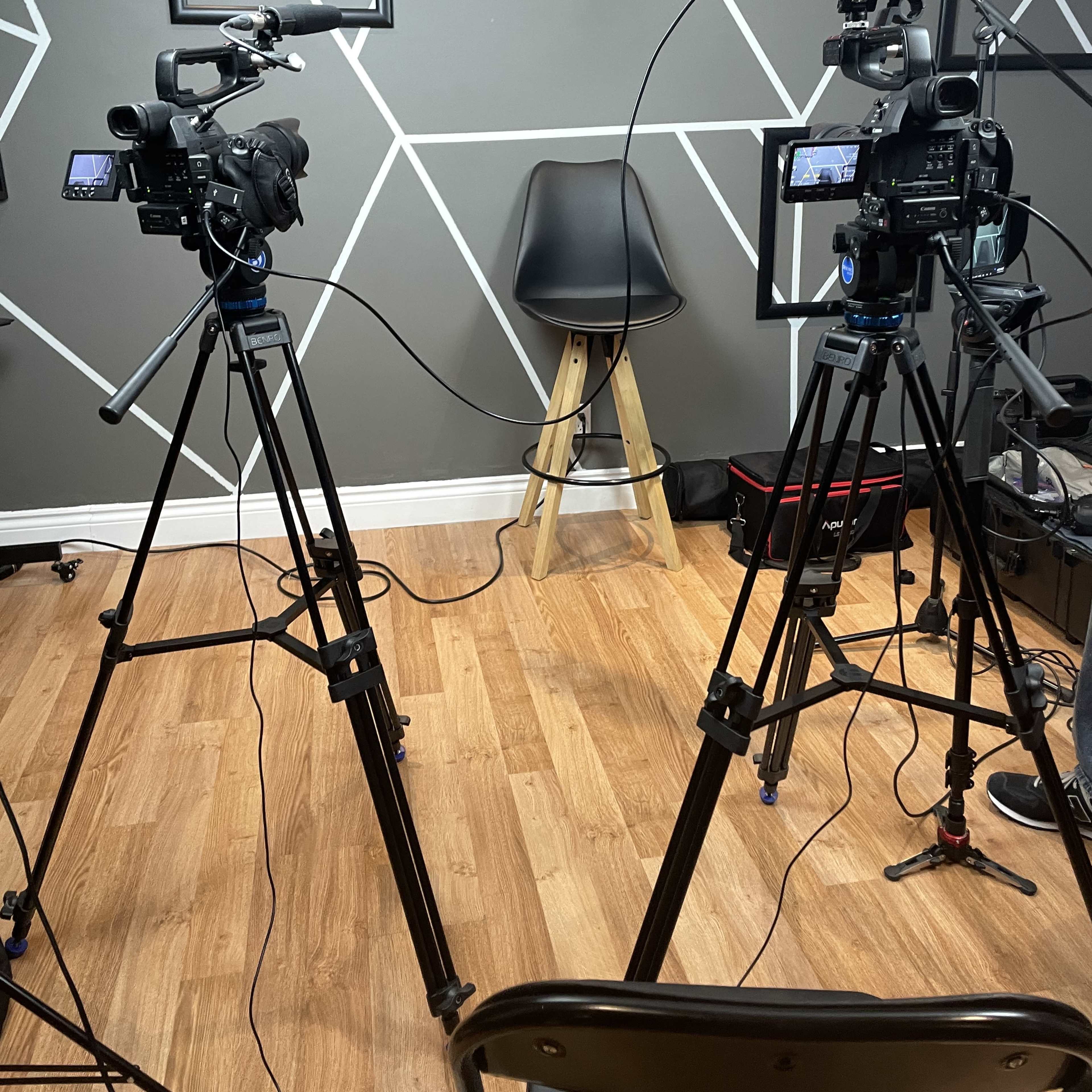 Two video cameras on tripods are positioned facing an empty chair in a studio with patterned walls.