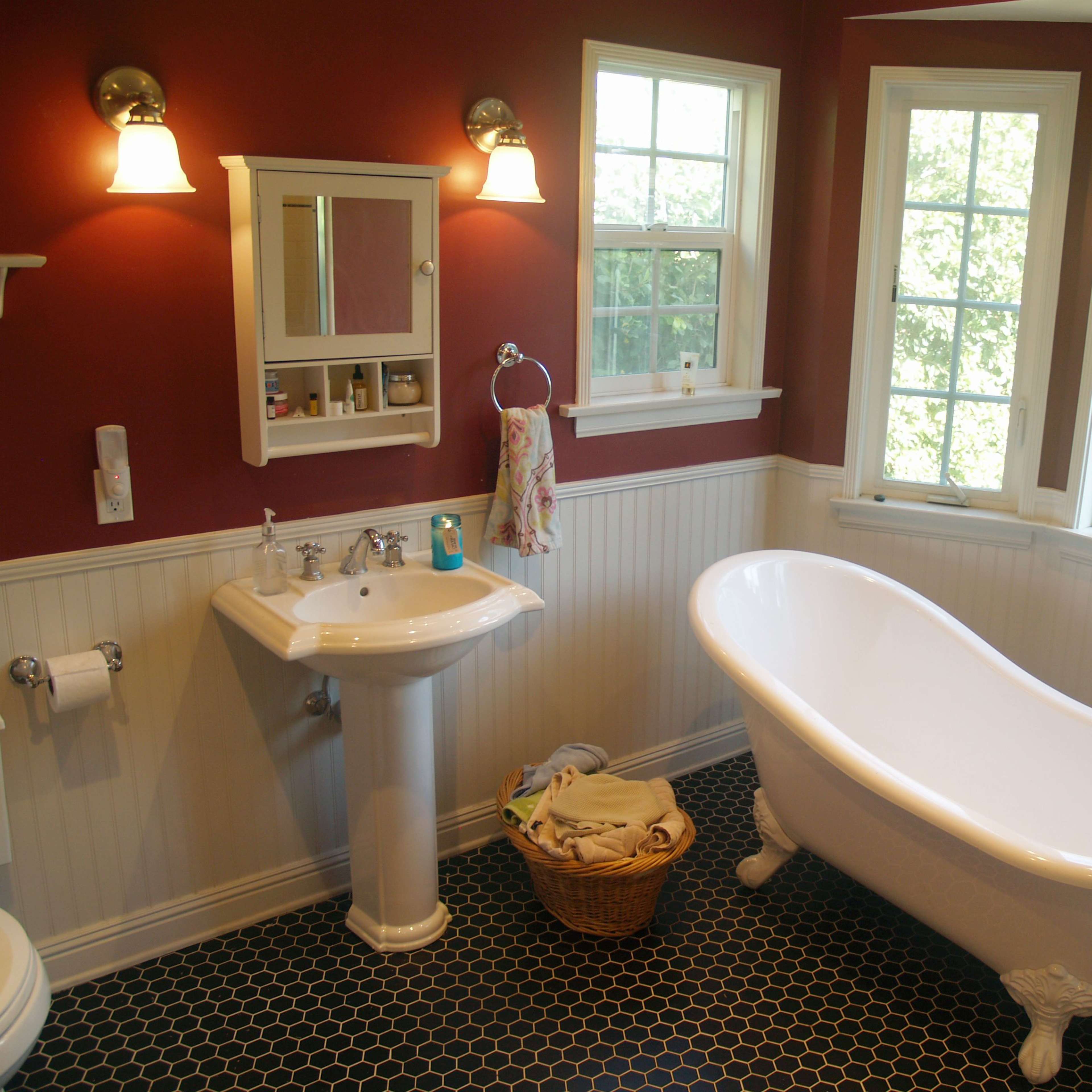A bathroom featuring a freestanding white bathtub, a pedestal sink, and large windows allowing natural light to enter, with a dark hexagonal tile floor.