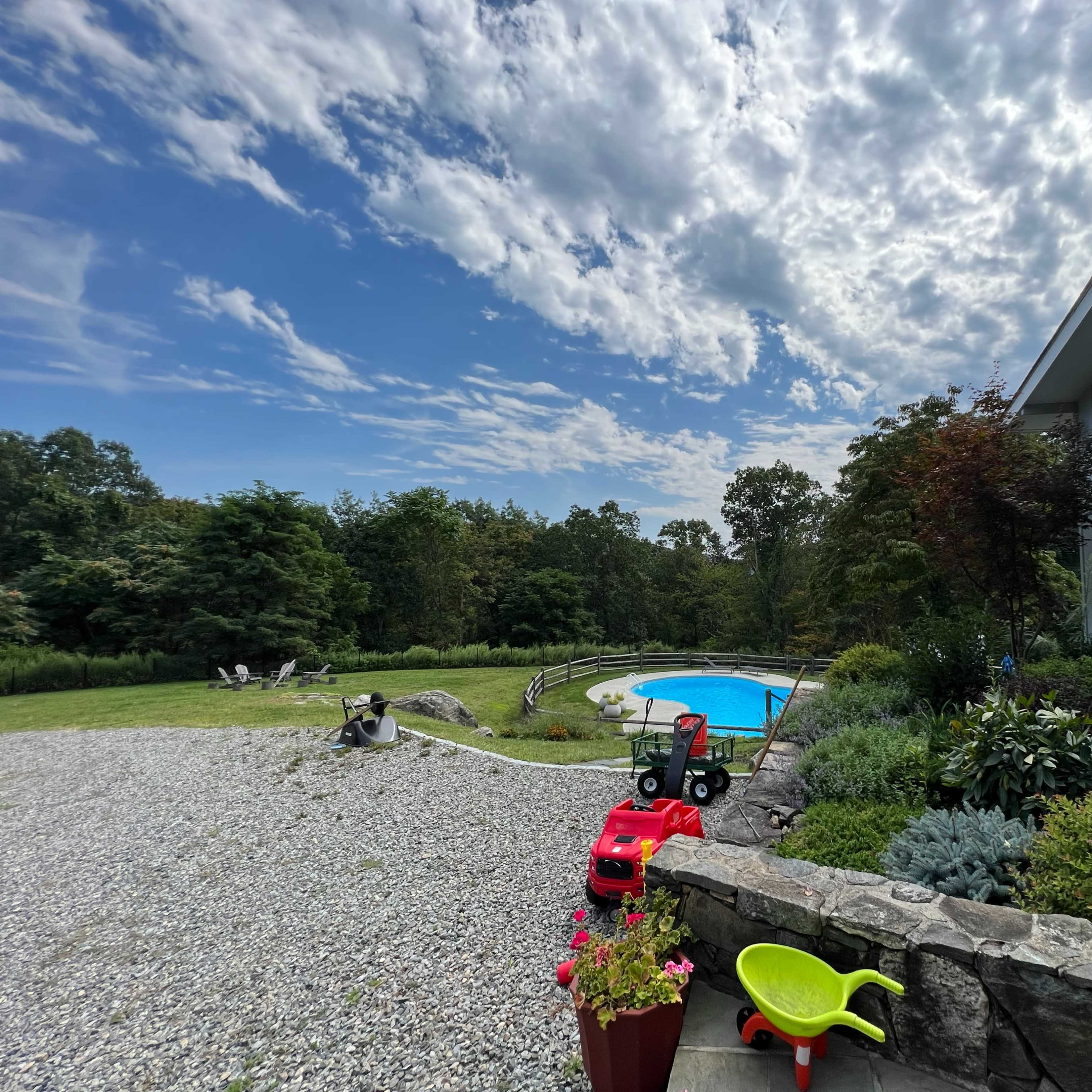 A landscaped backyard features a swimming pool surrounded by greenery, with a gravel area and a colorful toy truck parked nearby.