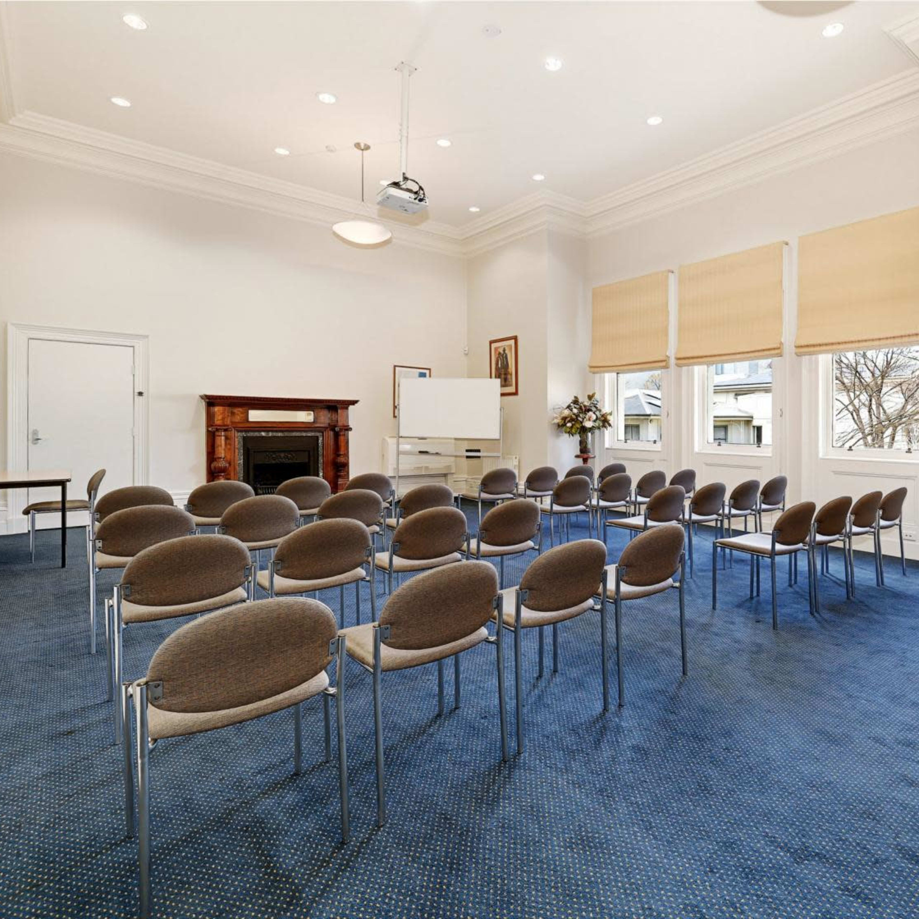 A meeting room with arranged chairs facing a presentation area, featuring a fireplace and large windows with shades.