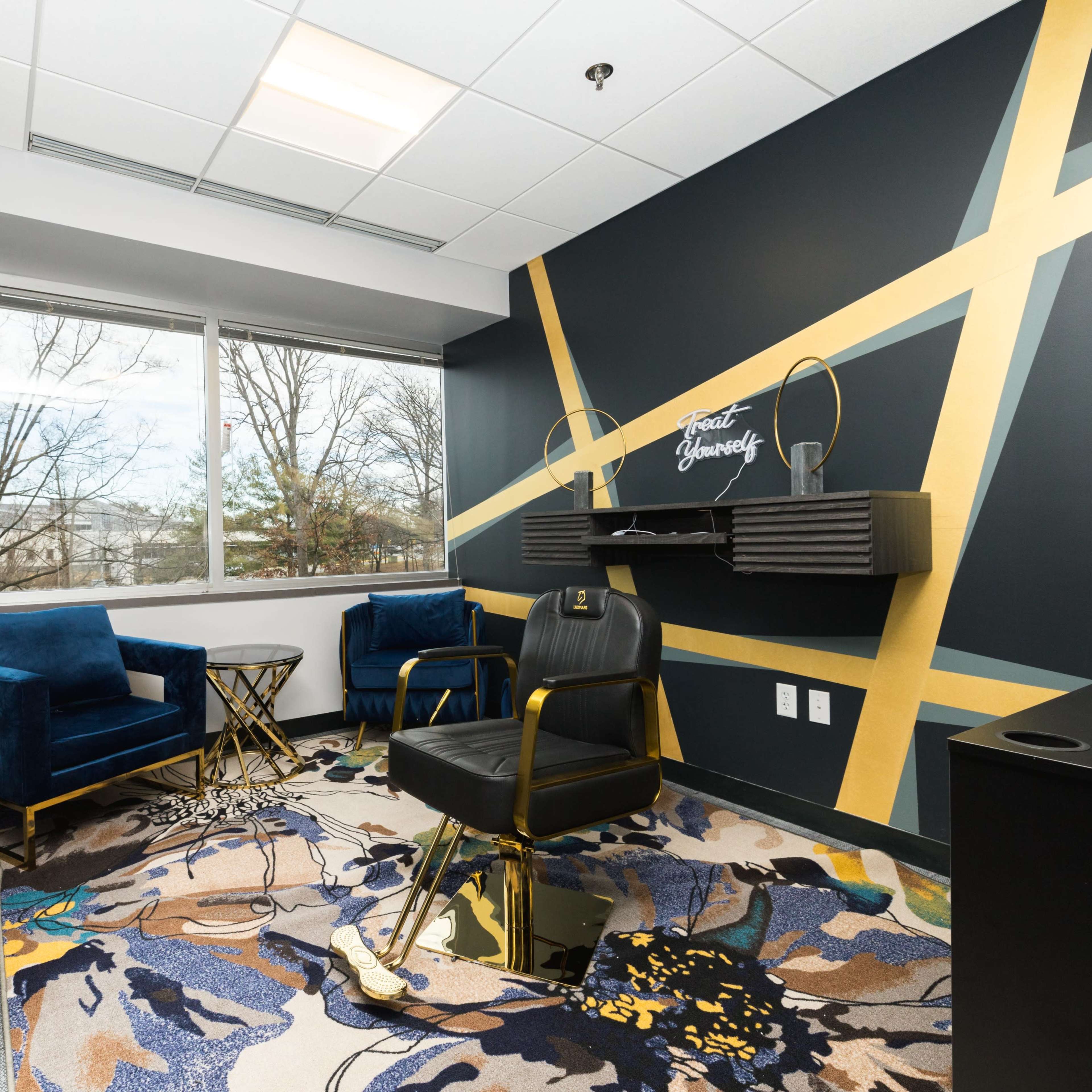 A modern office space featuring two blue armchairs, a black chair, and a decorative rug, with large windows allowing natural light and a minimalist black and gold color scheme on the walls.