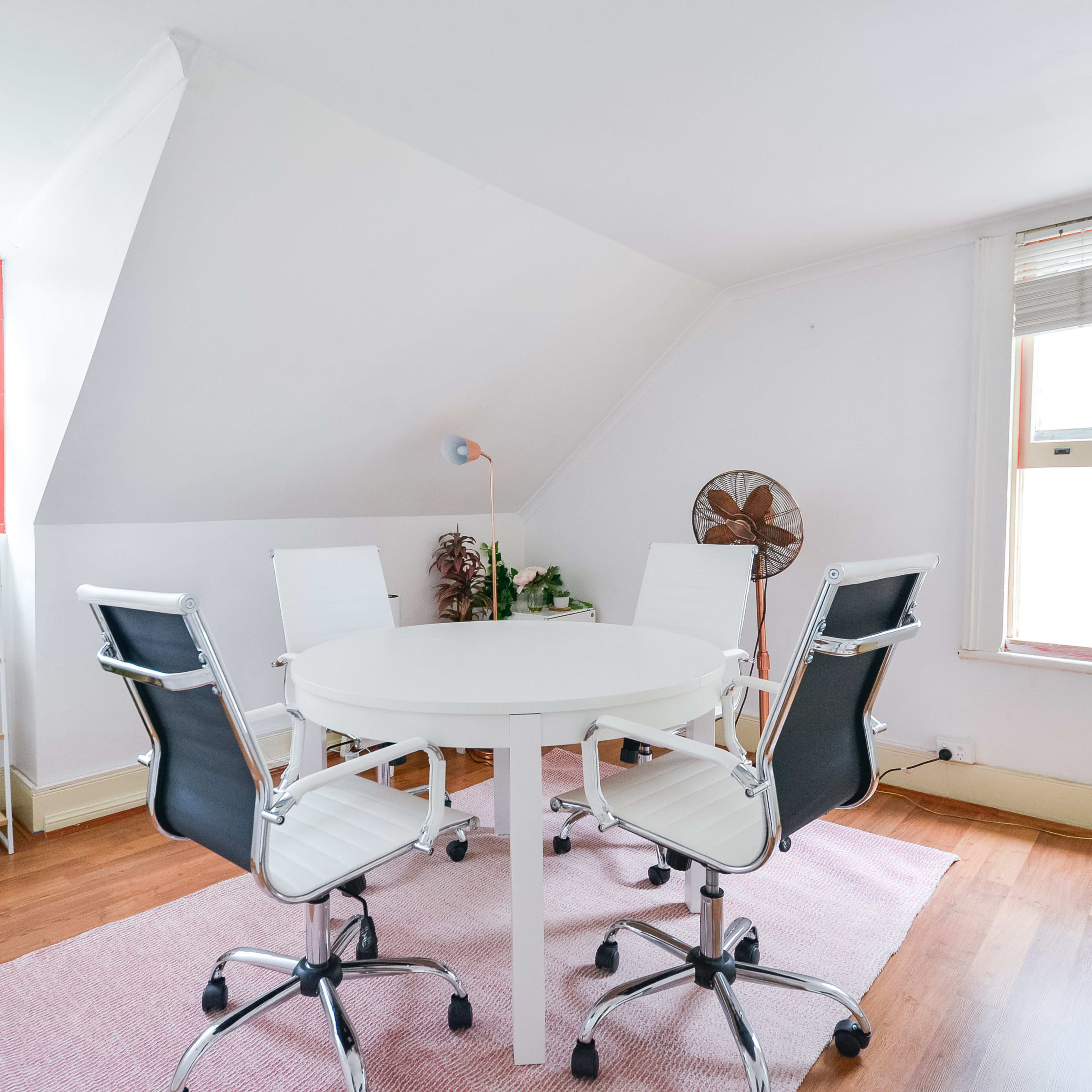 A small, bright room with a white round table surrounded by four black and white chairs, featuring a fan and a plant shelf against the walls.