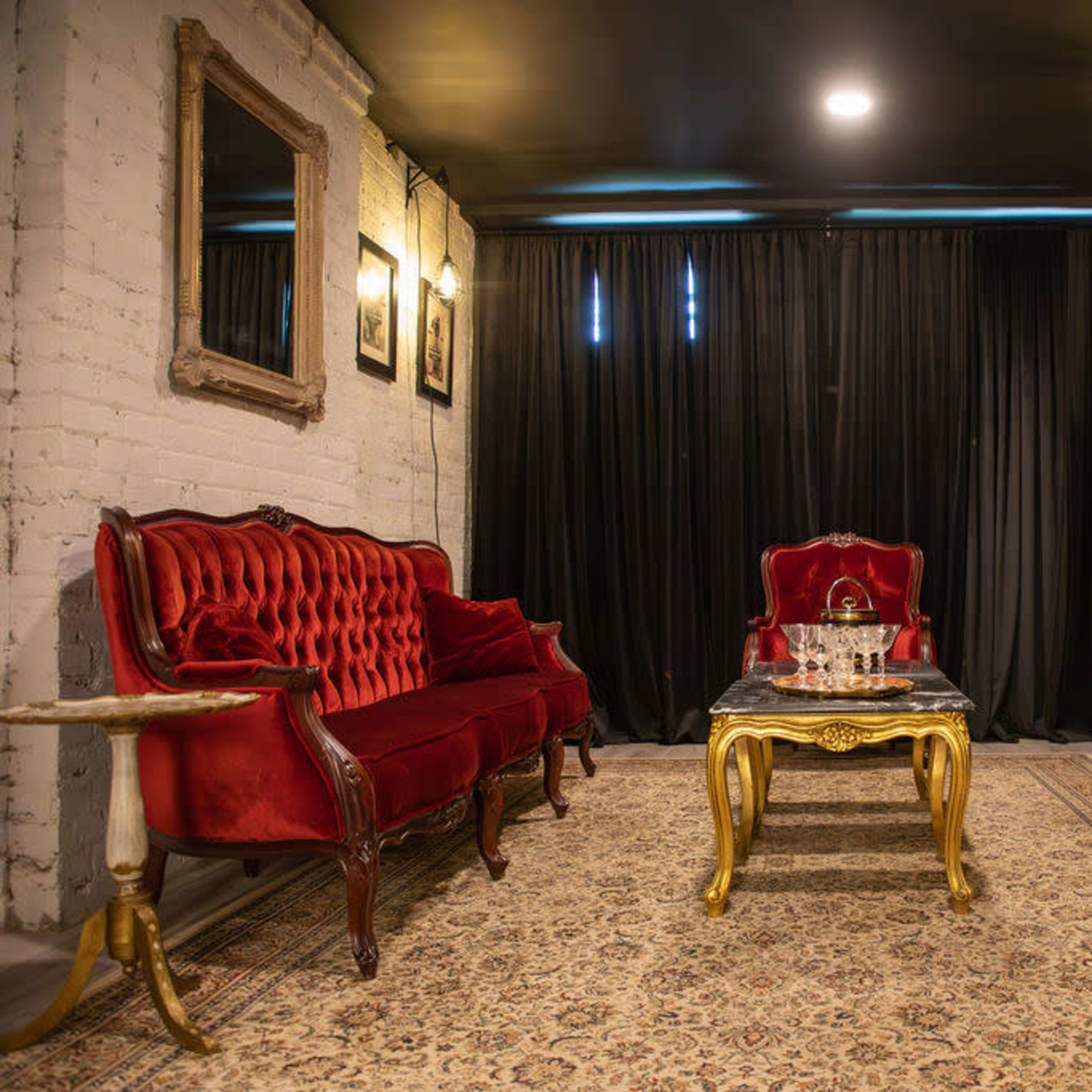 A sitting area with two red velvet sofas and a gold-accented coffee table on an ornate rug, surrounded by black curtains and framed mirrors on the walls.