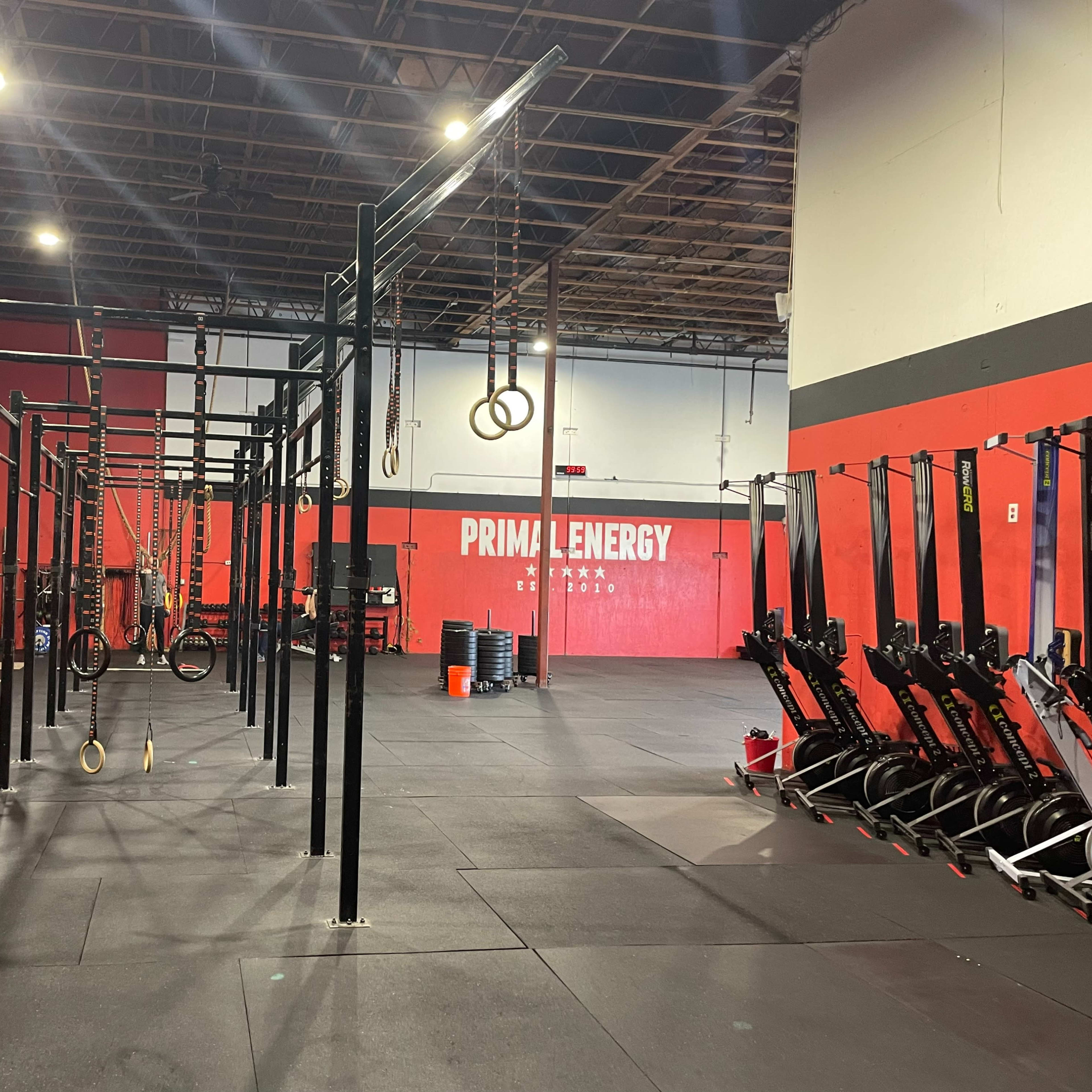 A gym interior with various fitness equipment, including pull-up bars, gymnastic rings, and rowing machines, against a backdrop of red and black walls.