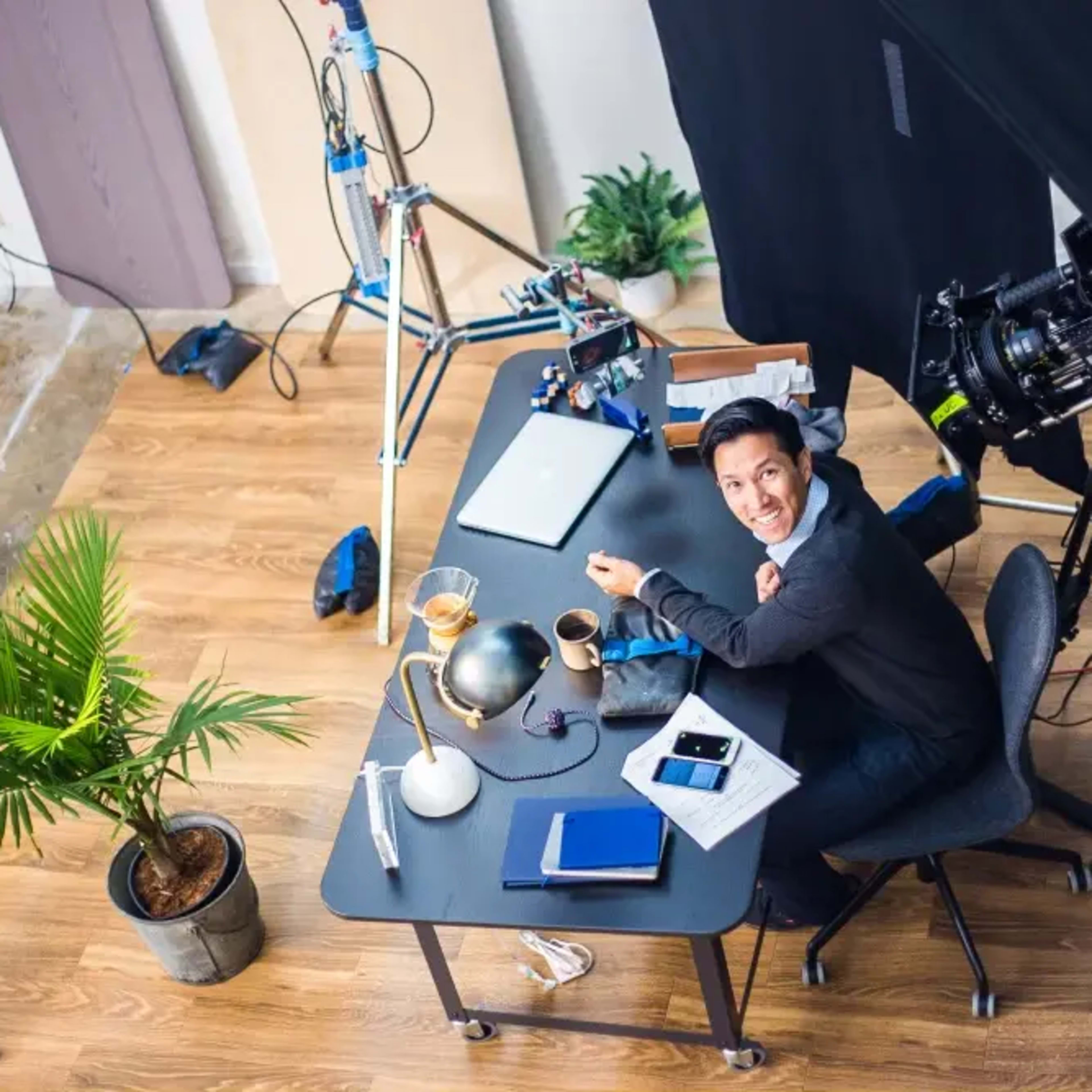 A person sits at a desk in a studio setup with a camera and lighting equipment nearby.