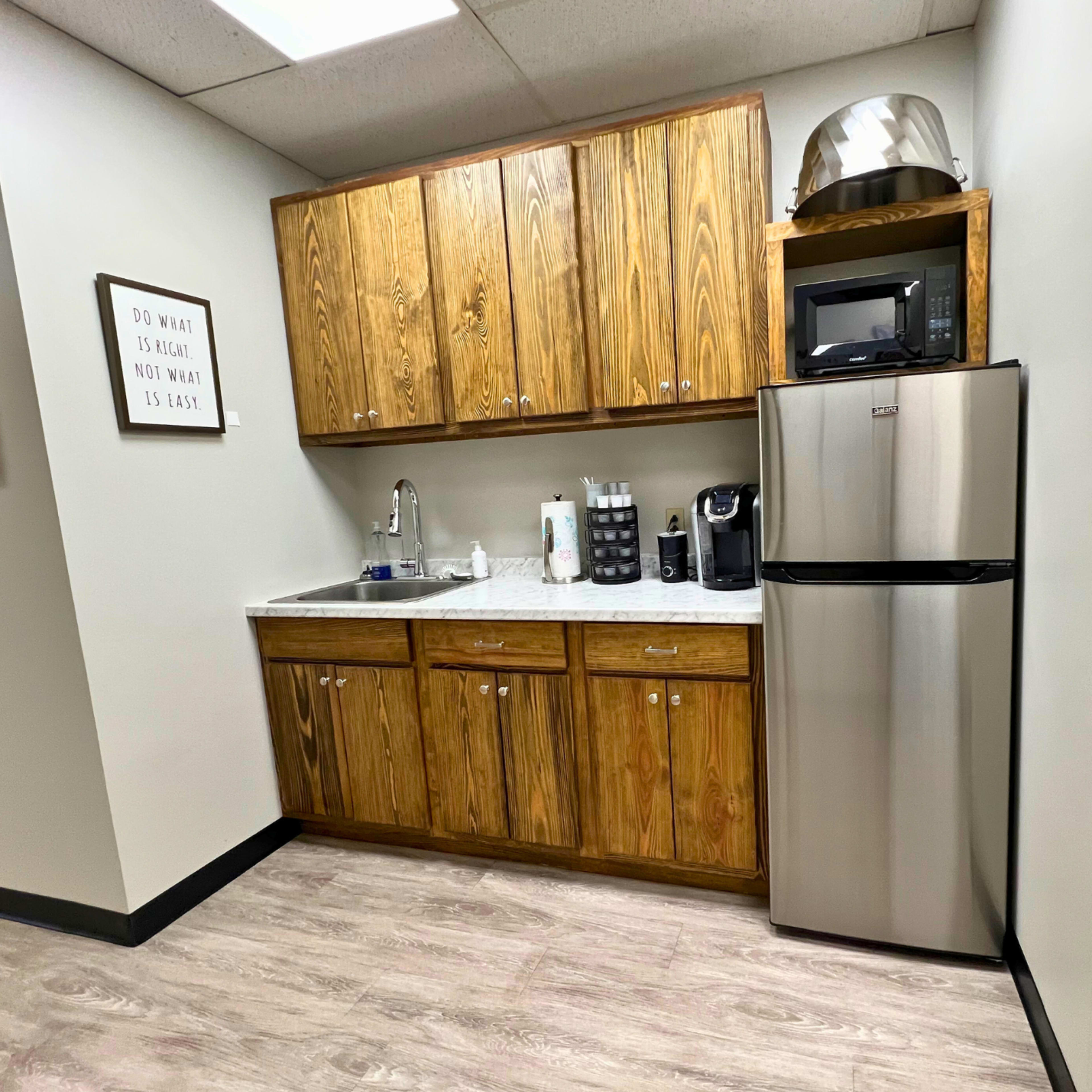 A small kitchen area with wooden cabinets, a countertop, a microwave, a coffee maker, and a stainless steel refrigerator.
