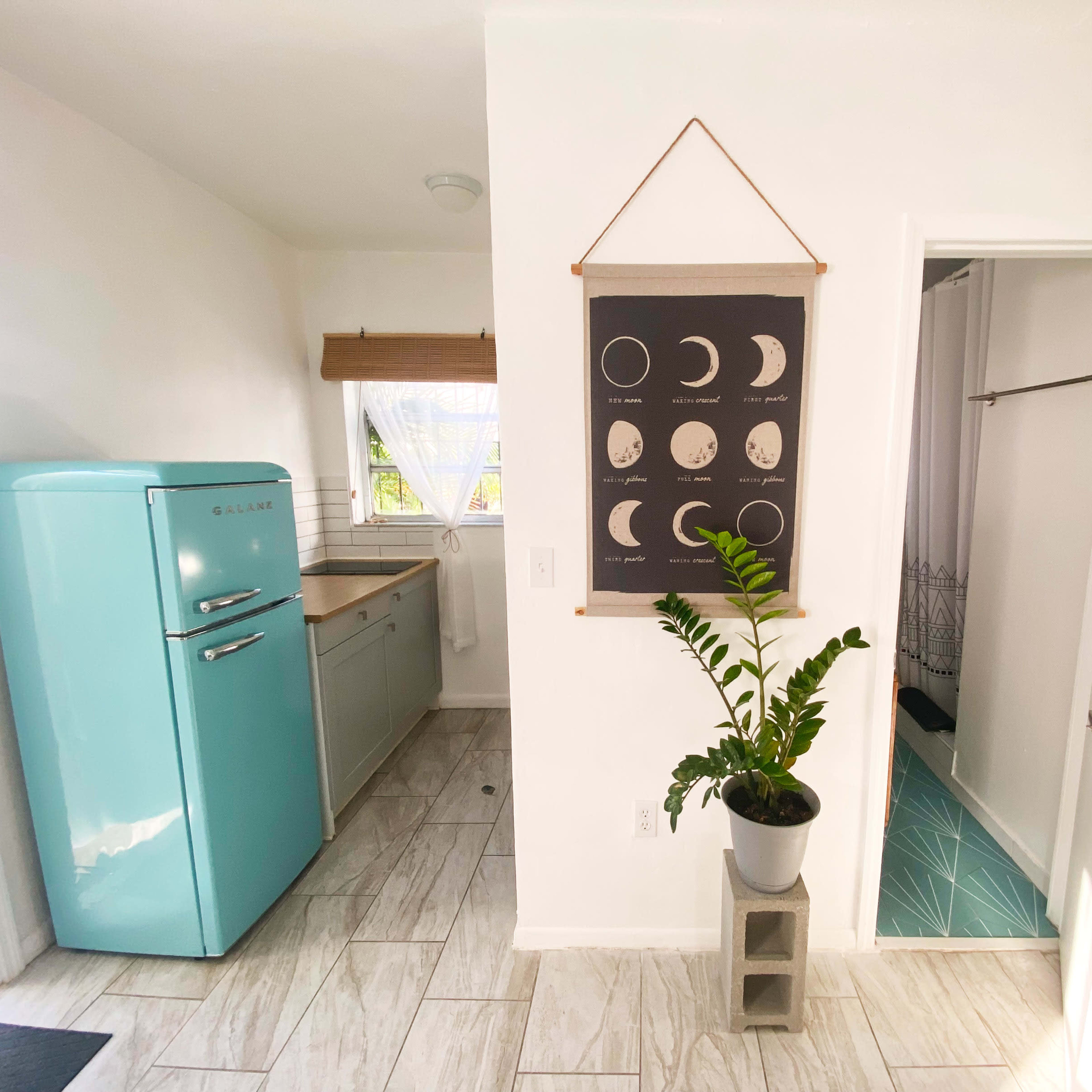 A bright, minimalist kitchen area featuring a turquoise refrigerator, a plant in a pot, and a wall hanging displaying phases of the moon.