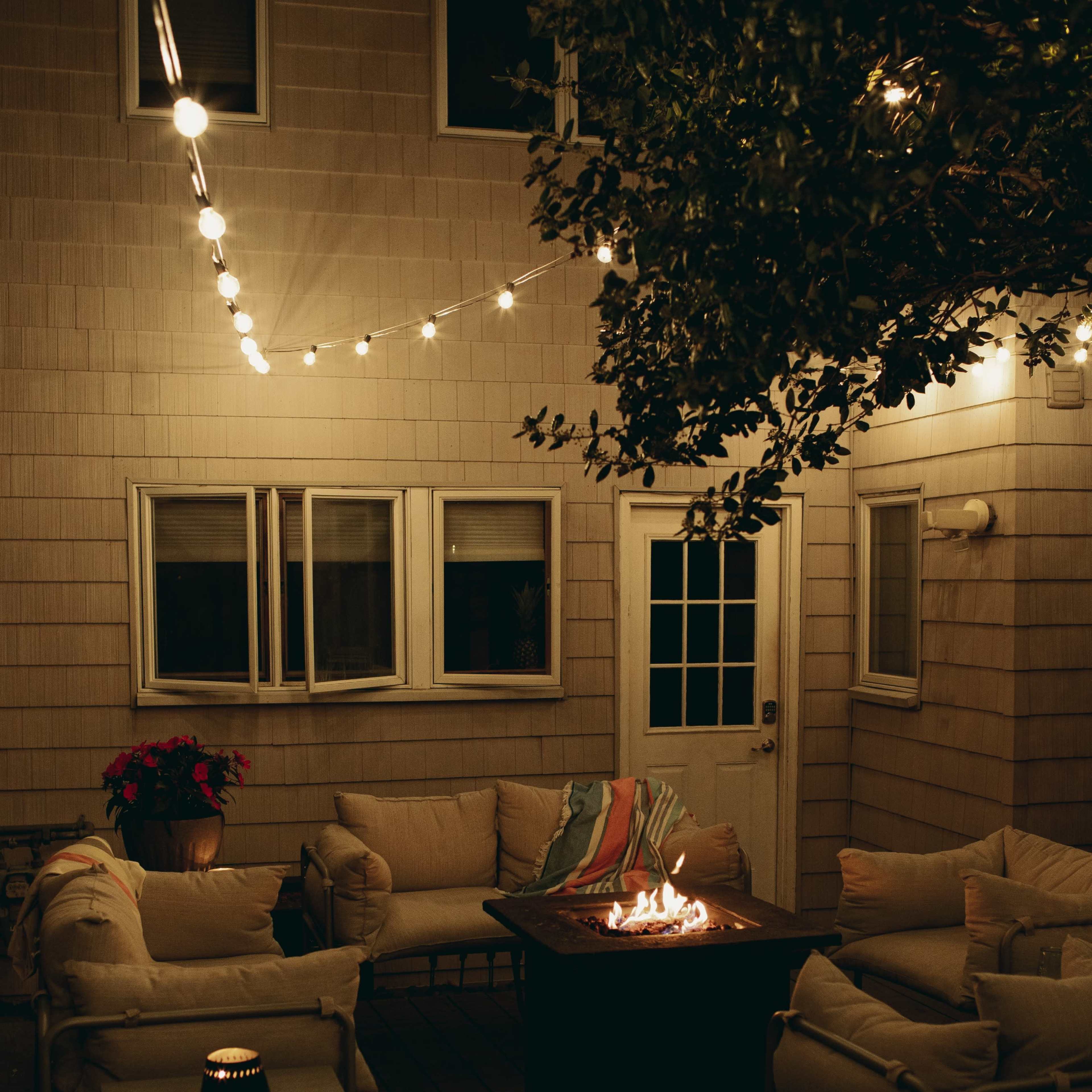 A cozy outdoor seating area is illuminated by string lights, featuring a fire pit surrounded by cushioned chairs.